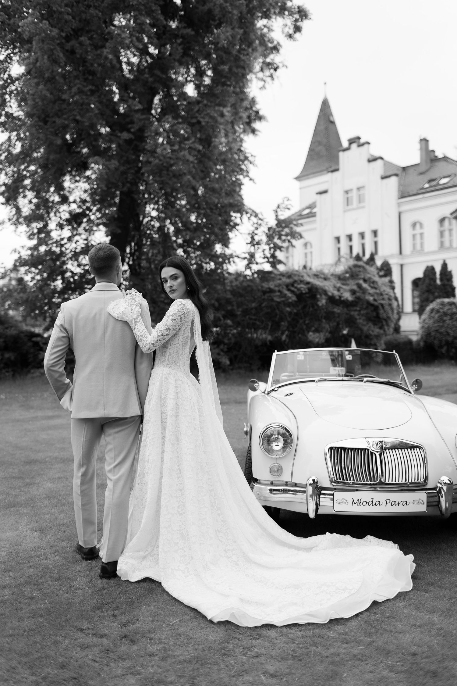 Black and white photo of bride and groom standing by a vintage convertible car outside an elegant mansion.