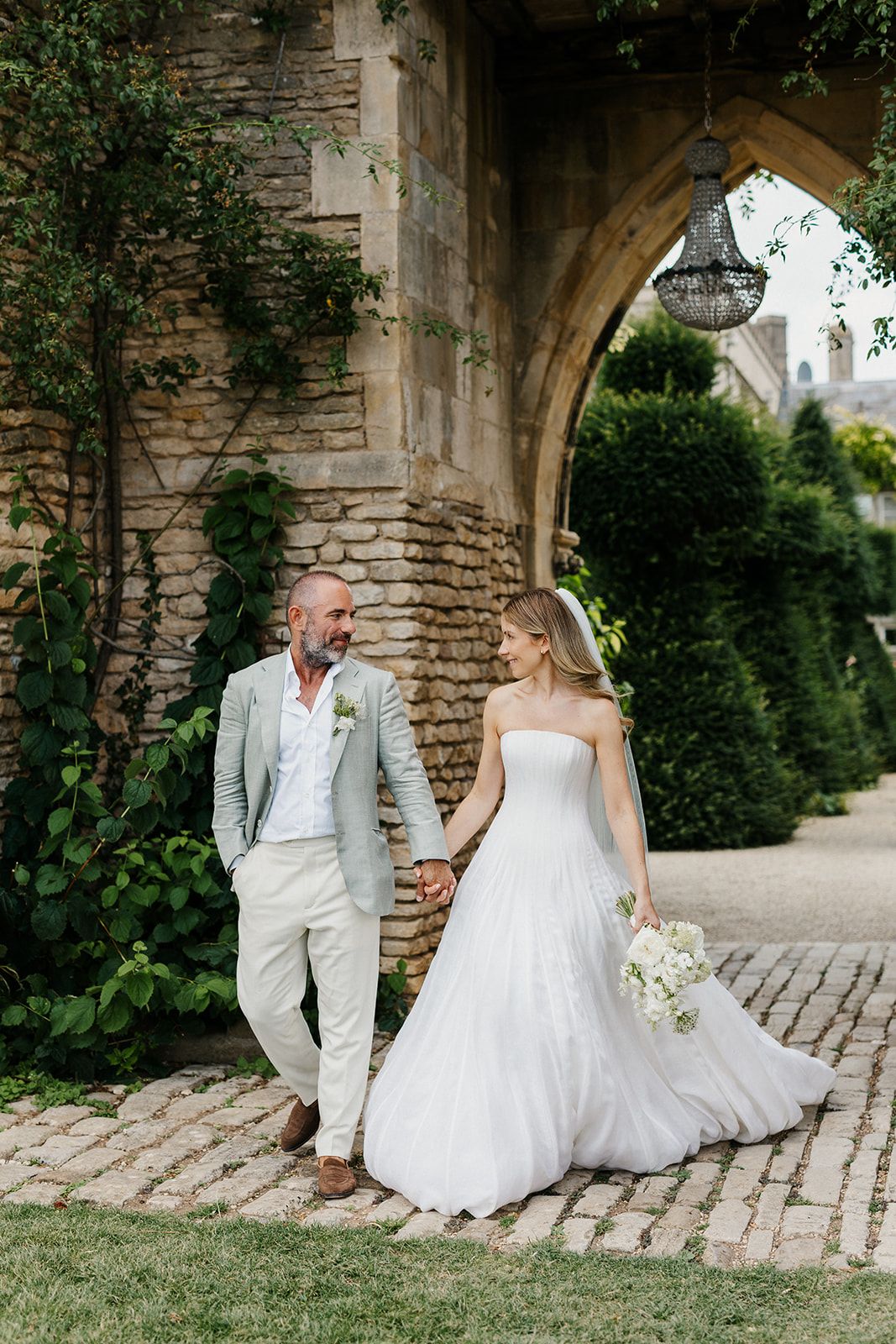 Bride and groom walk hand-in-hand on cobblestone path beside ivy-covered stone arch at elegant outdoor wedding venue.
