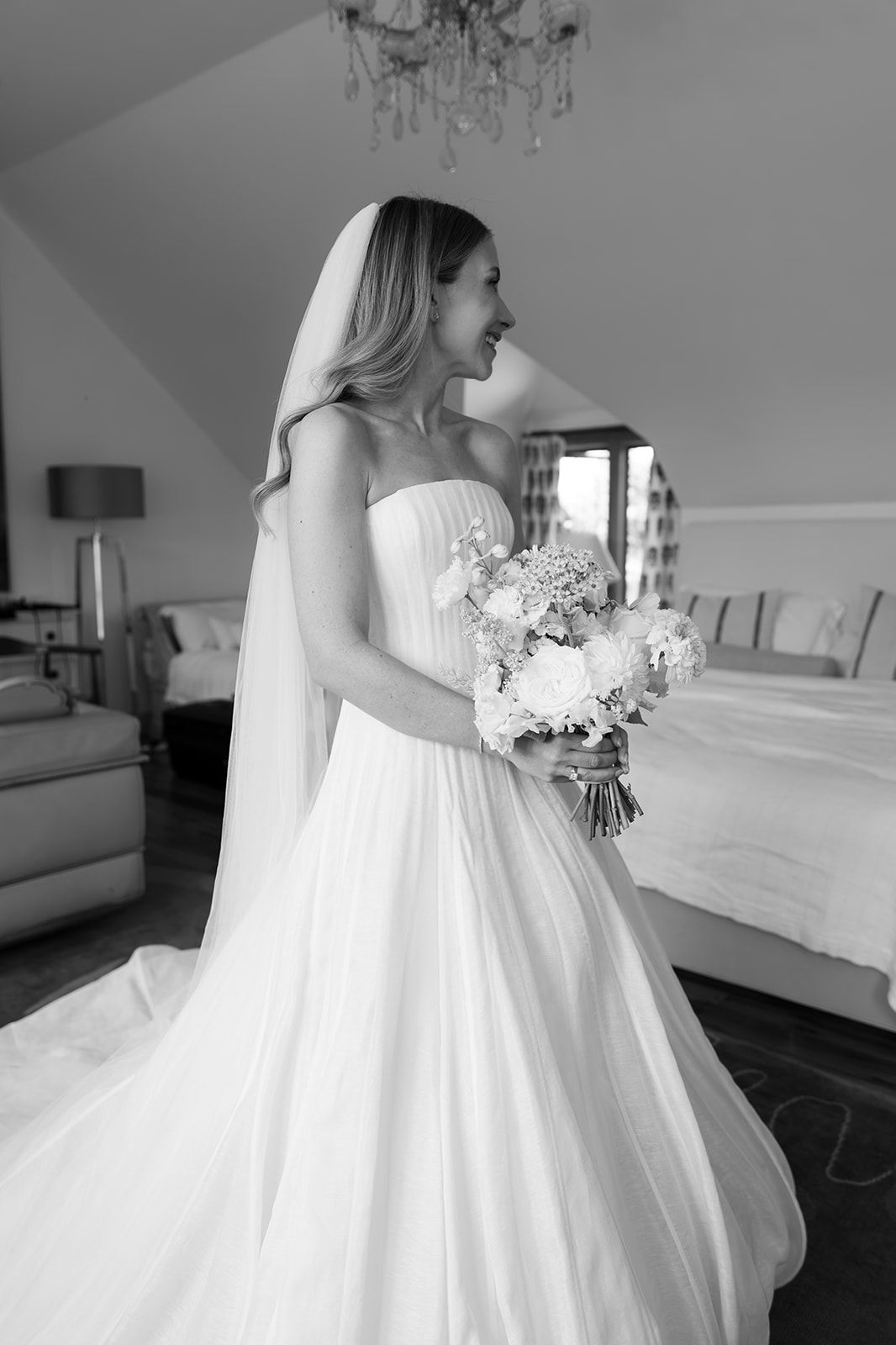 Bride in strapless white gown and veil holds floral bouquet in elegant room, black and white wedding photo.