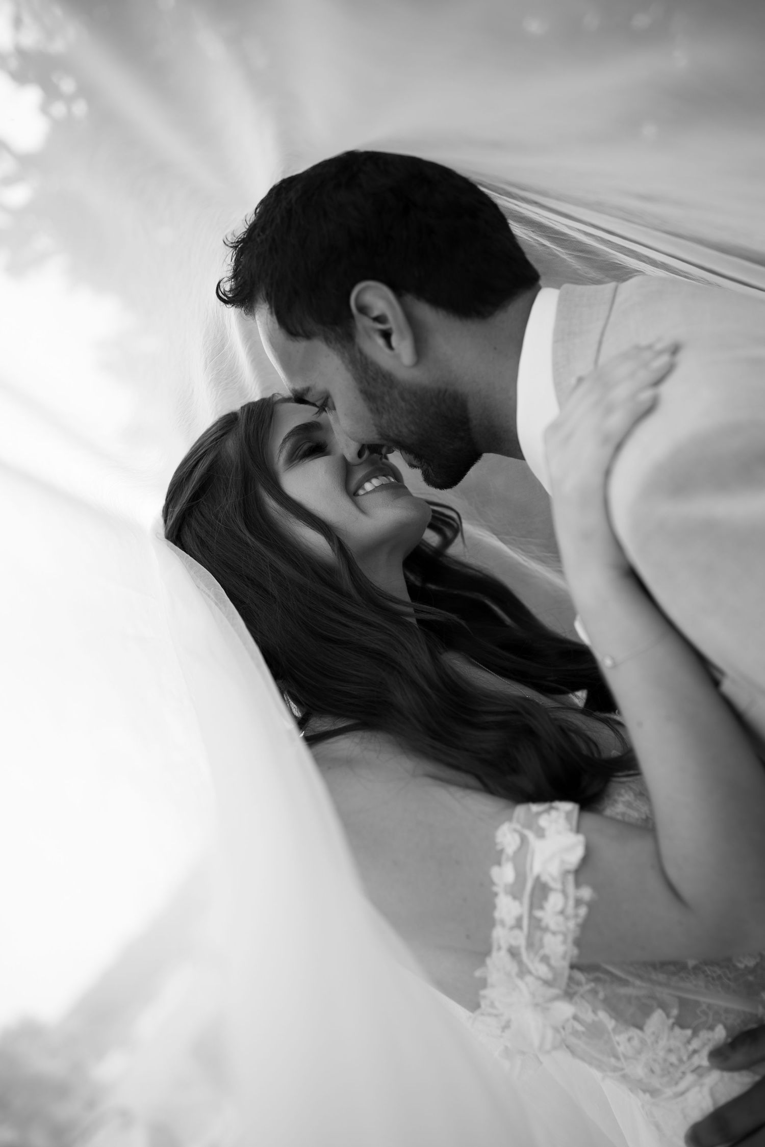 Black and white wedding photo of bride and groom sharing romantic moment under flowing veil.