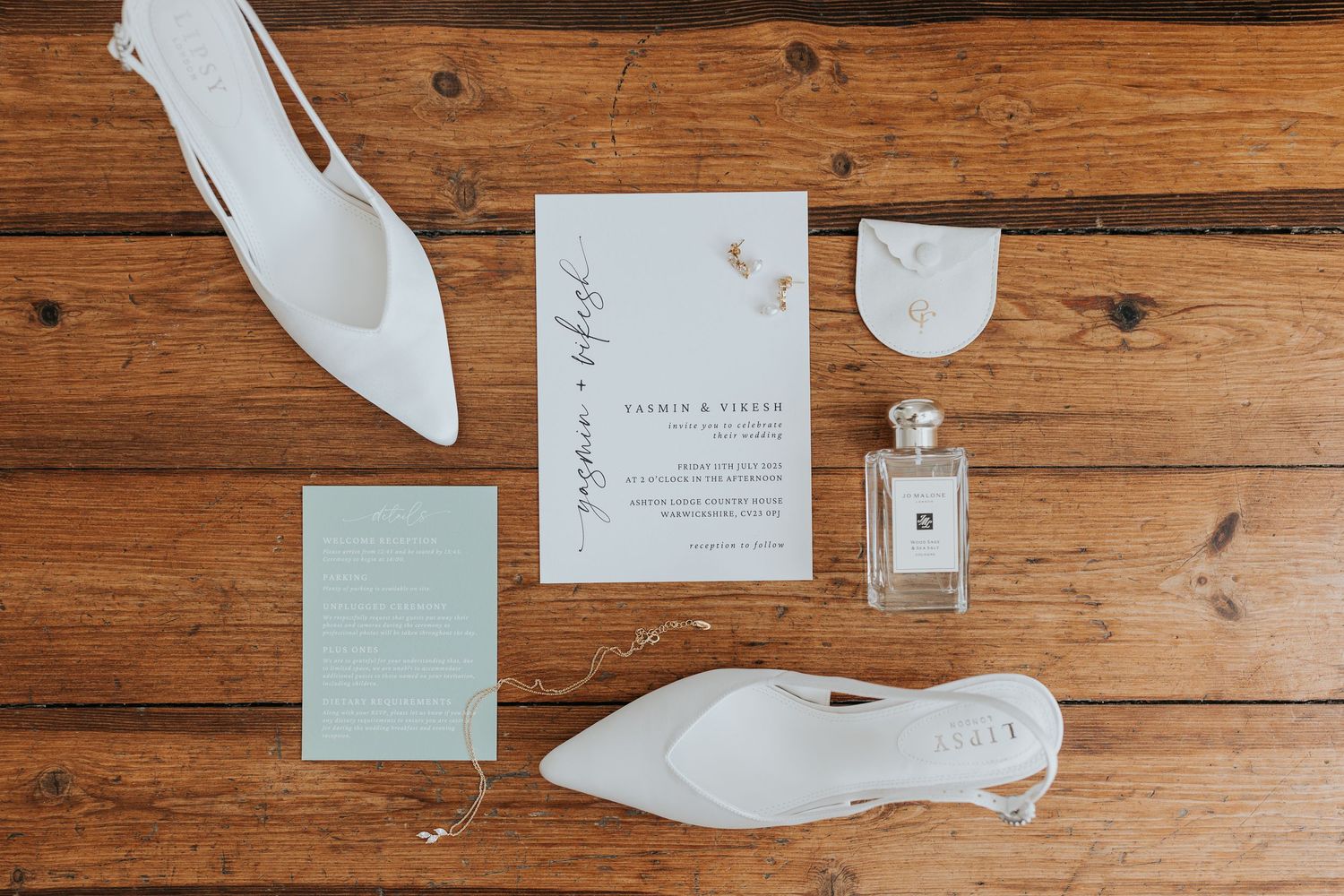 Elegant wedding flat lay featuring white heels, Jo Malone perfume, floral invitations, and stationery on rustic wood floor.