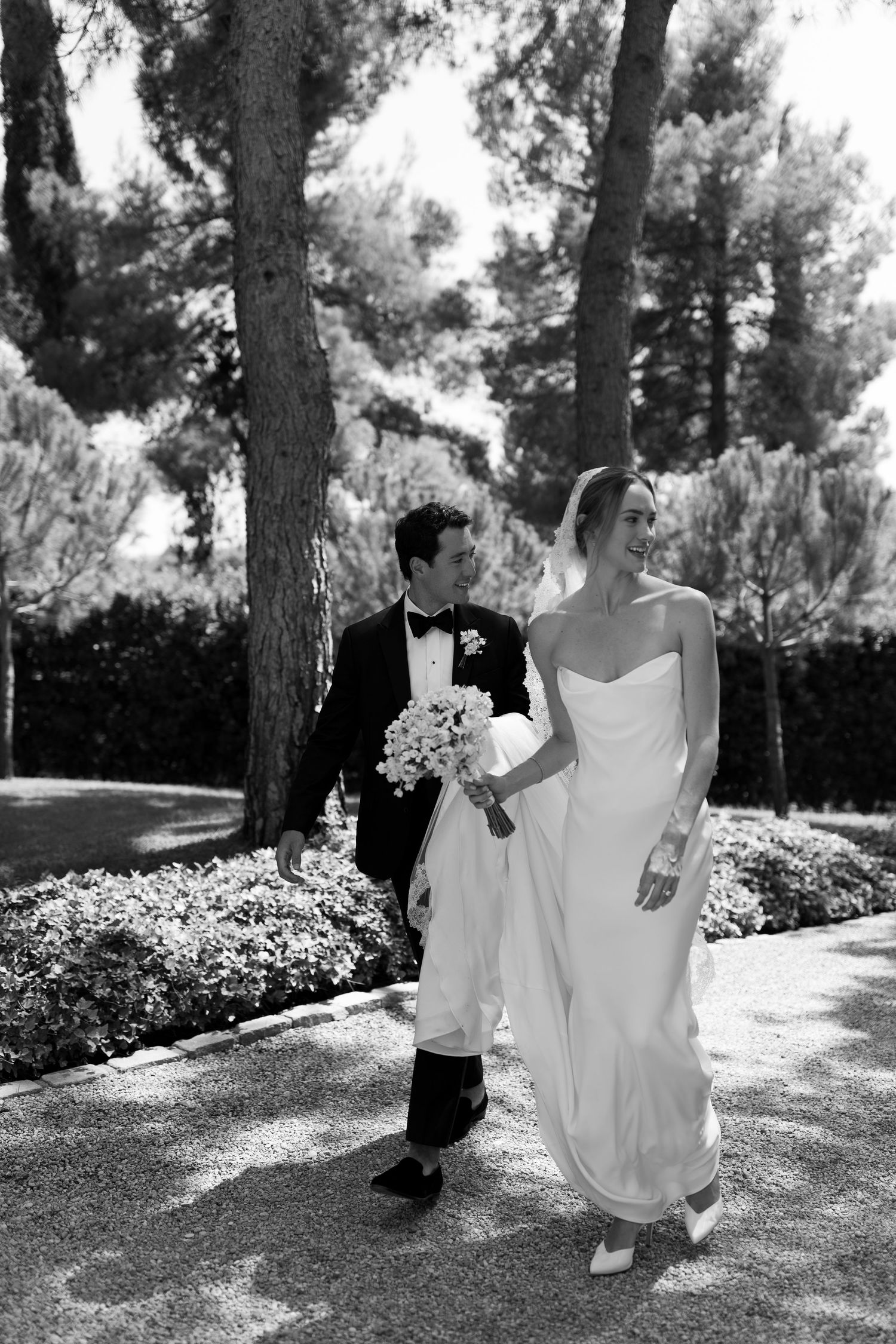 Black and white photo of a bride and groom walking together outdoors, surrounded by lush trees and greenery.