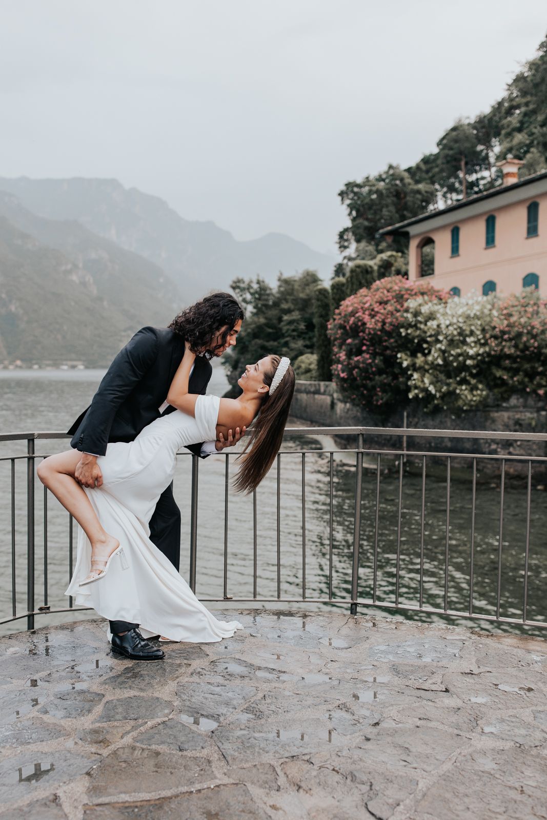 A newlywed couple embracing on a balcony overlooking a scenic lake and mountains in the background in Lake Como