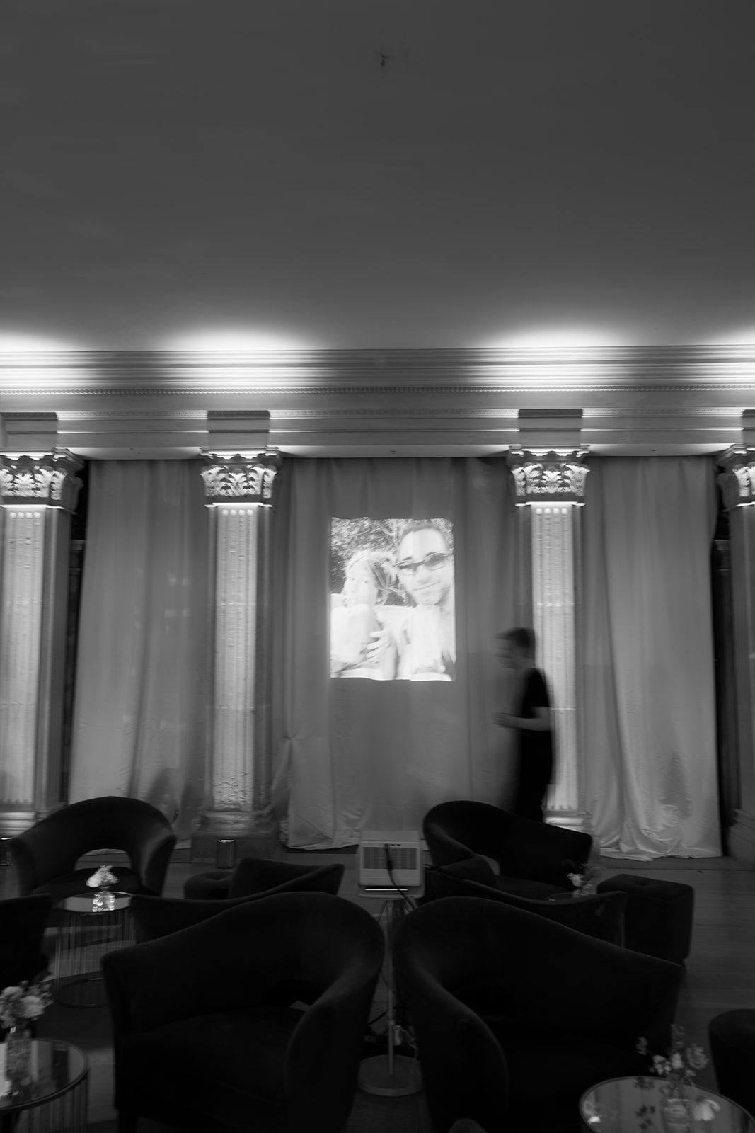 Black and white photo of an elegant lounge with columns, curtains, projected image, and blurred figure walking past seating area.
