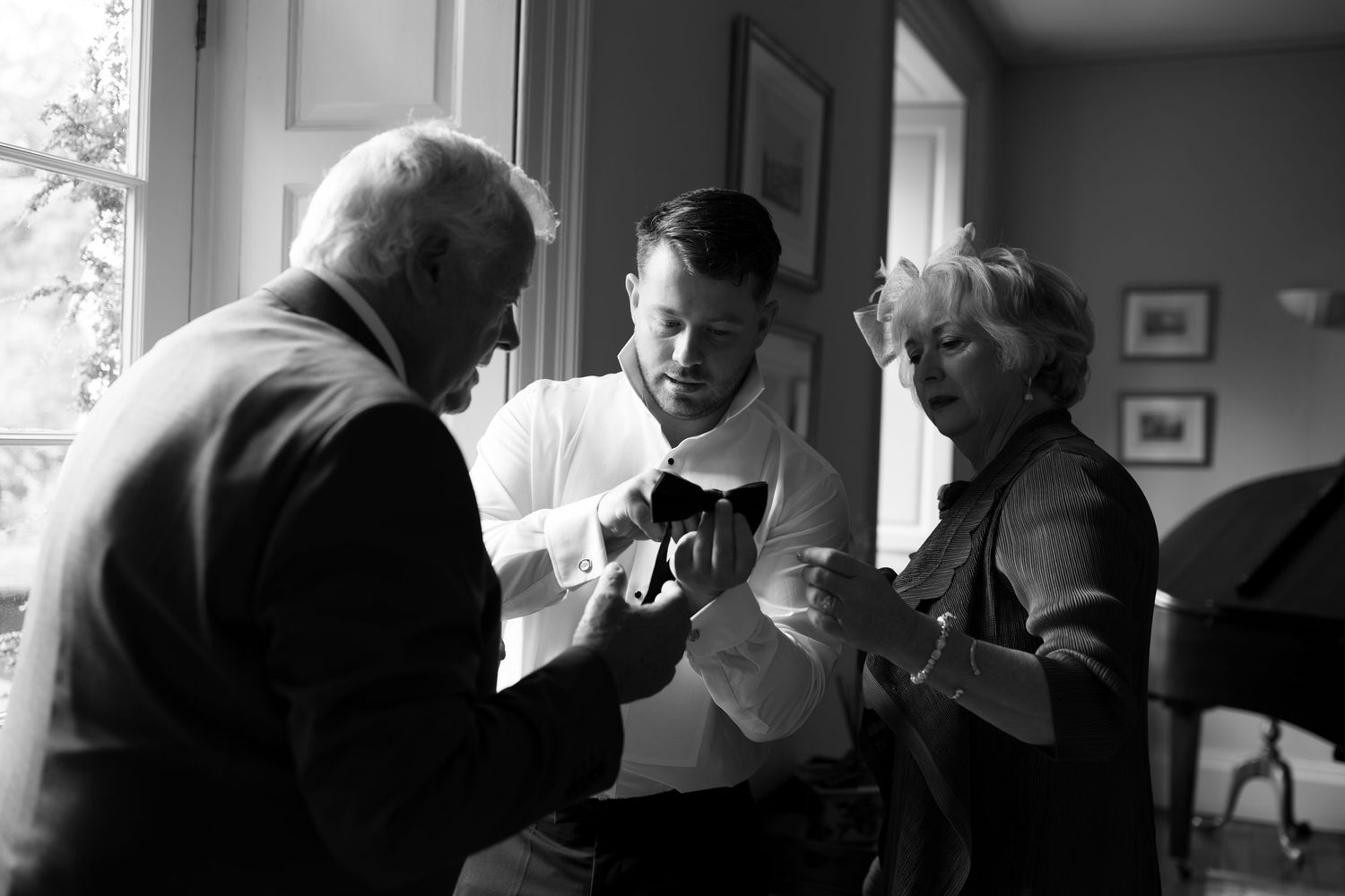 Black and white photo of three people examining an object together near a bright window indoors.
