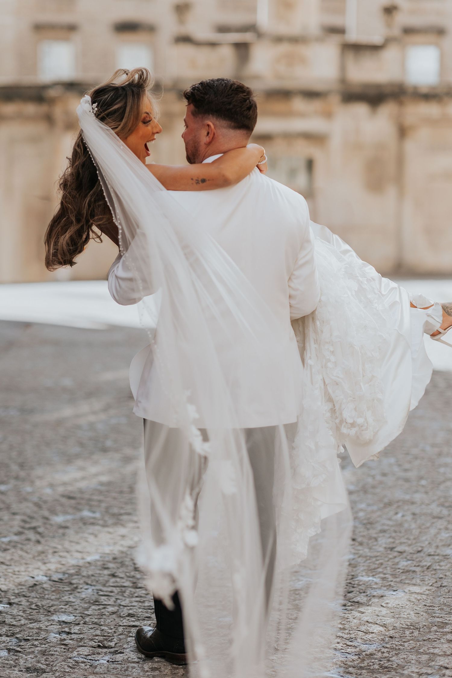 Groom in white suit carries bride in lace wedding dress and veil, sharing a romantic moment outdoors.