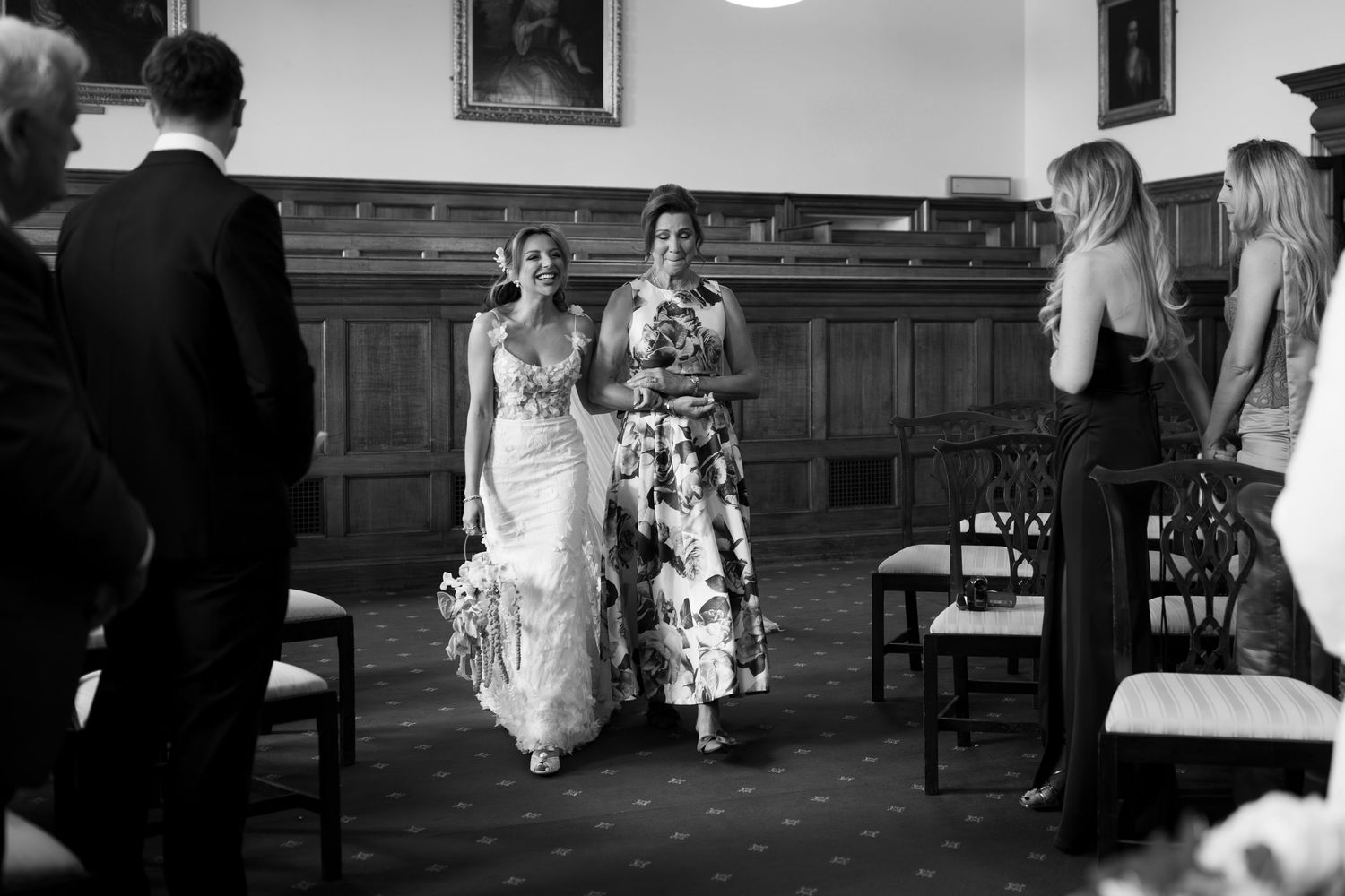 Black and white photo of two women walking down the aisle at an elegant wedding ceremony indoors.