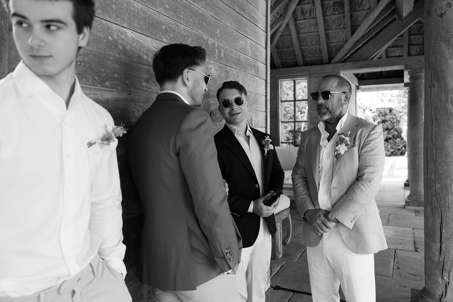Groomsmen in suits chat and laugh outside rustic barn venue in black and white wedding photo.