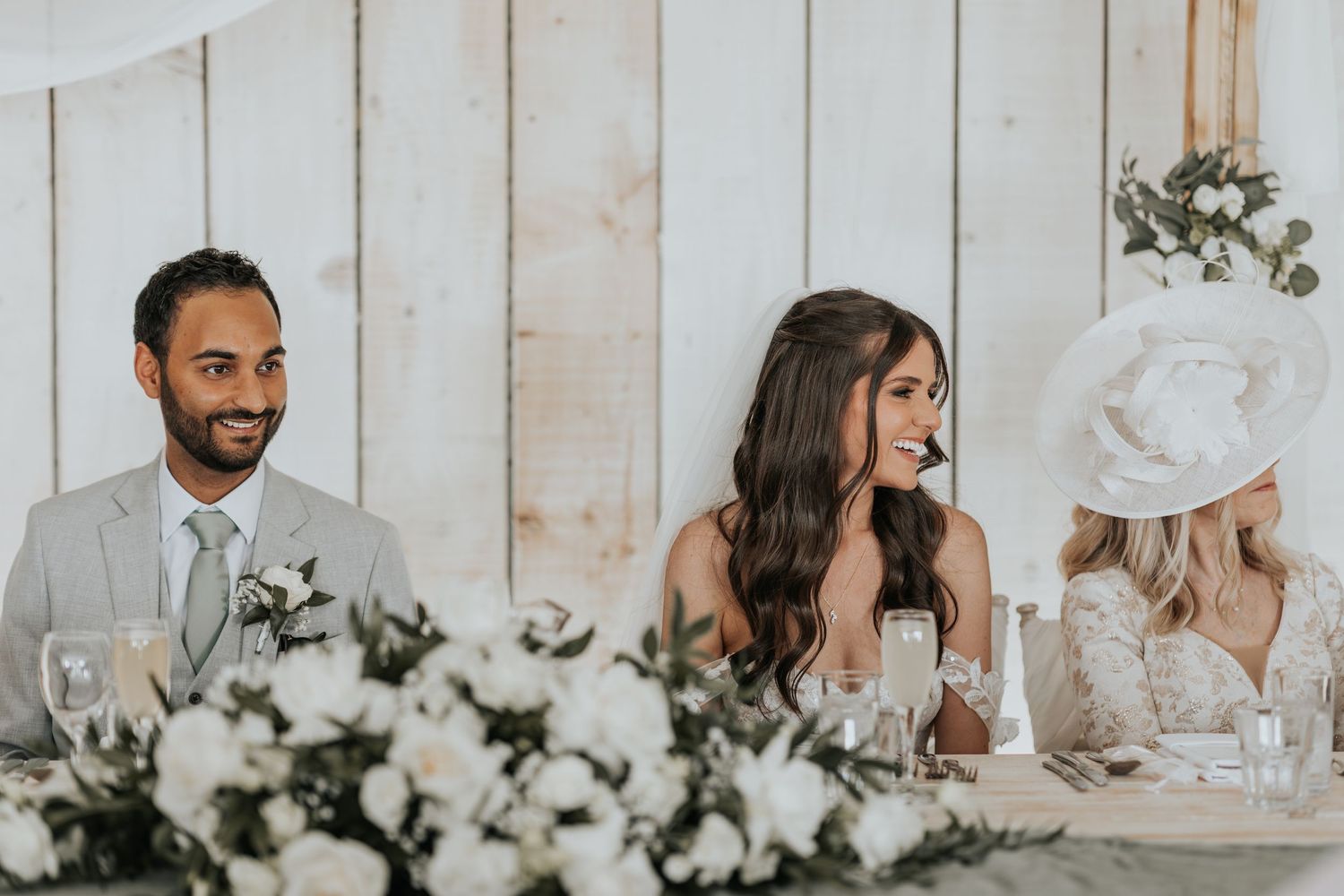 Smiling wedding guests seated at a decorated reception table with white floral arrangements in an elegant barn venue.