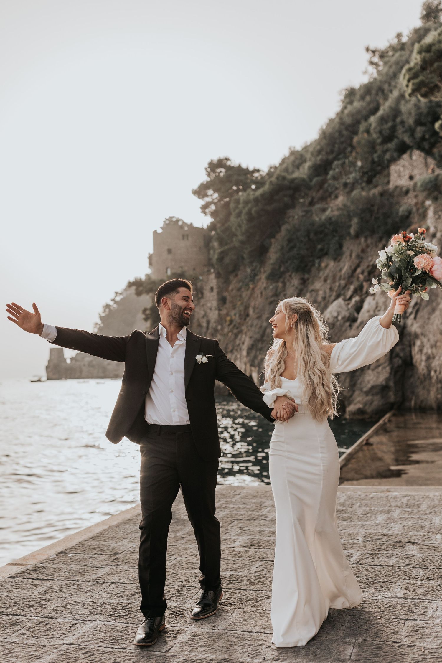 A couple dressed in formal attire walks along a rocky coastline, with mountains in the background in Positano