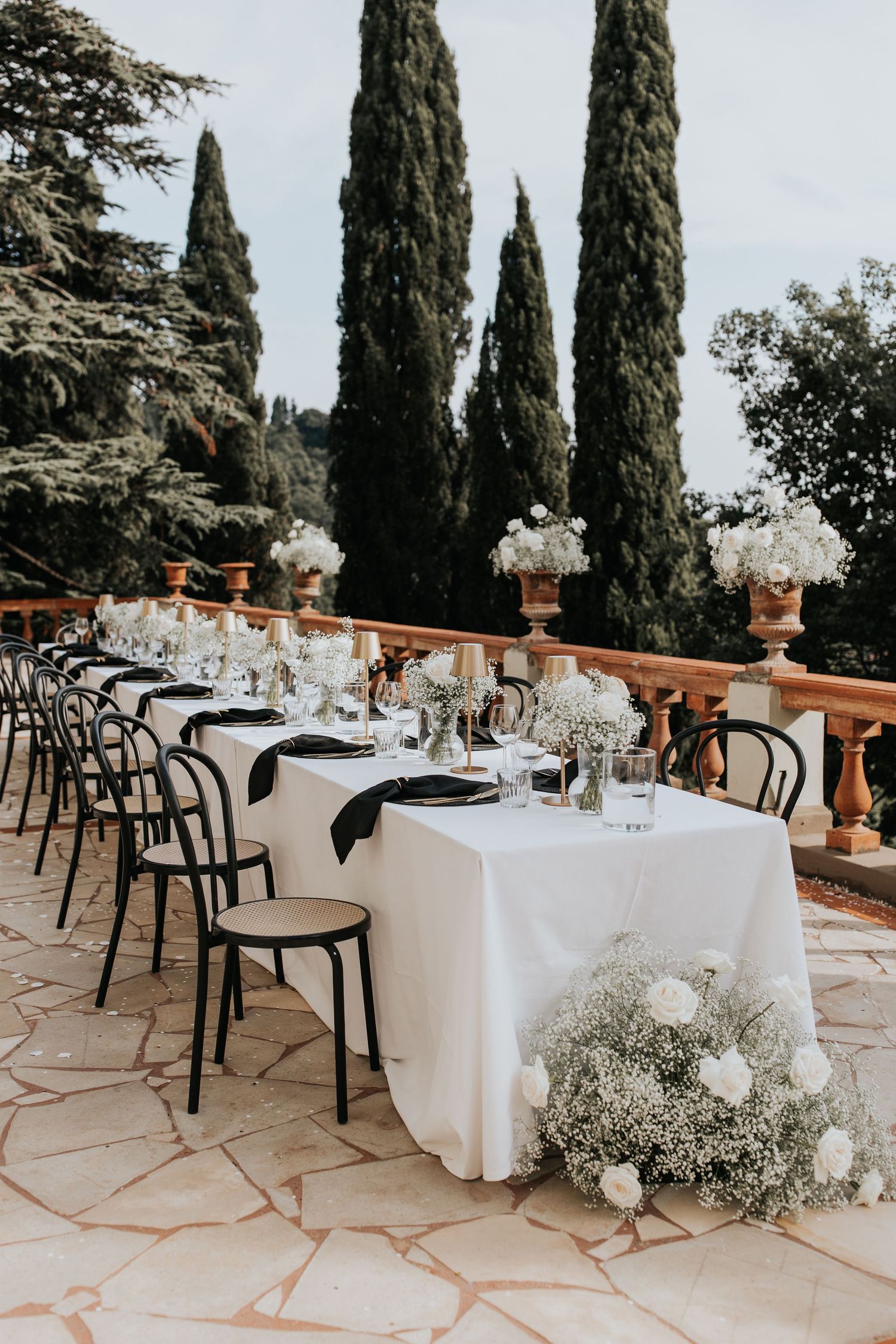 An outdoor dining area with white tablecloths, chairs, and a backdrop of tall cypress trees.