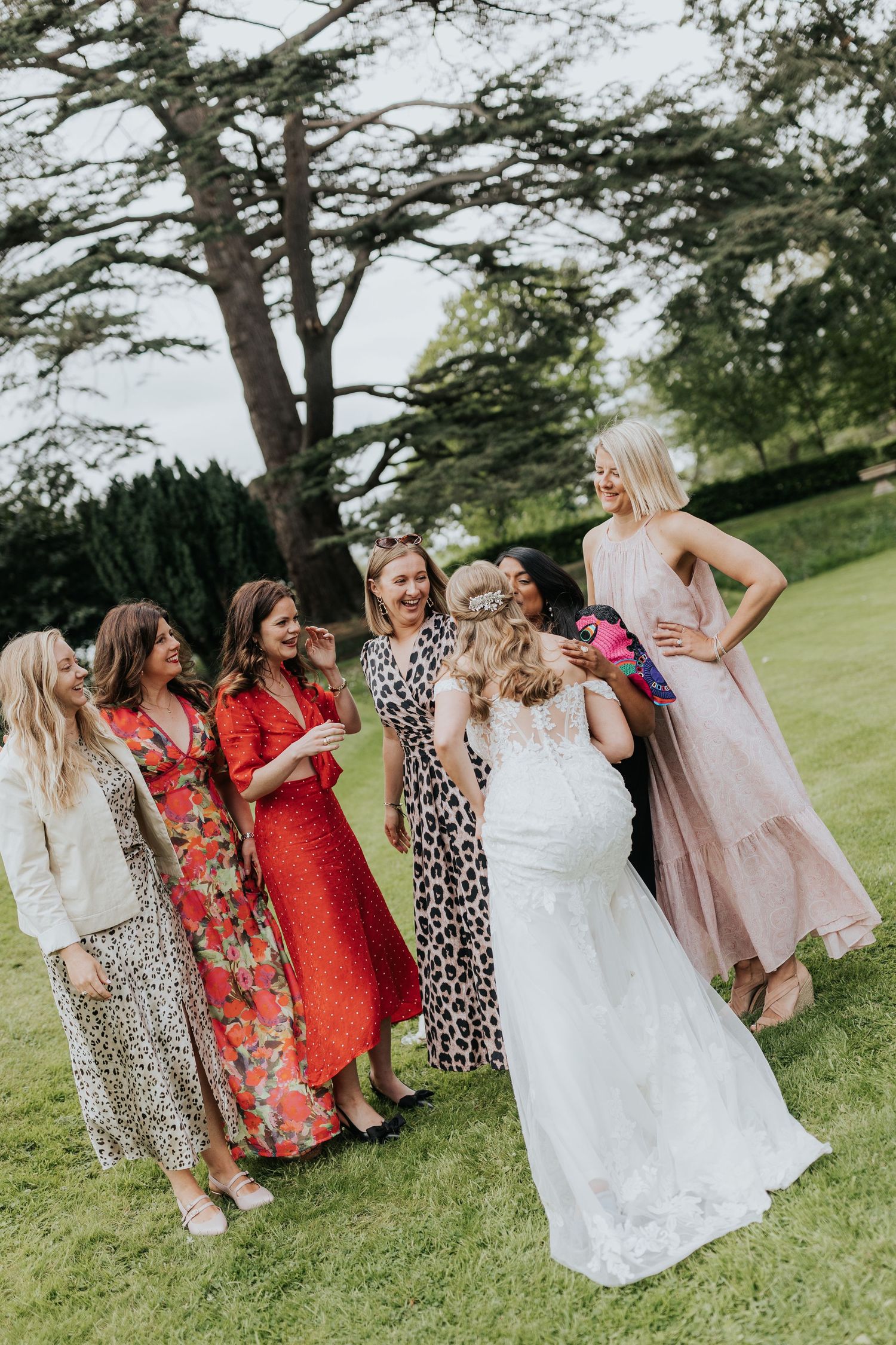 A bride in a white lace gown laughs with her bridesmaids in colorful dresses on a green lawn.
