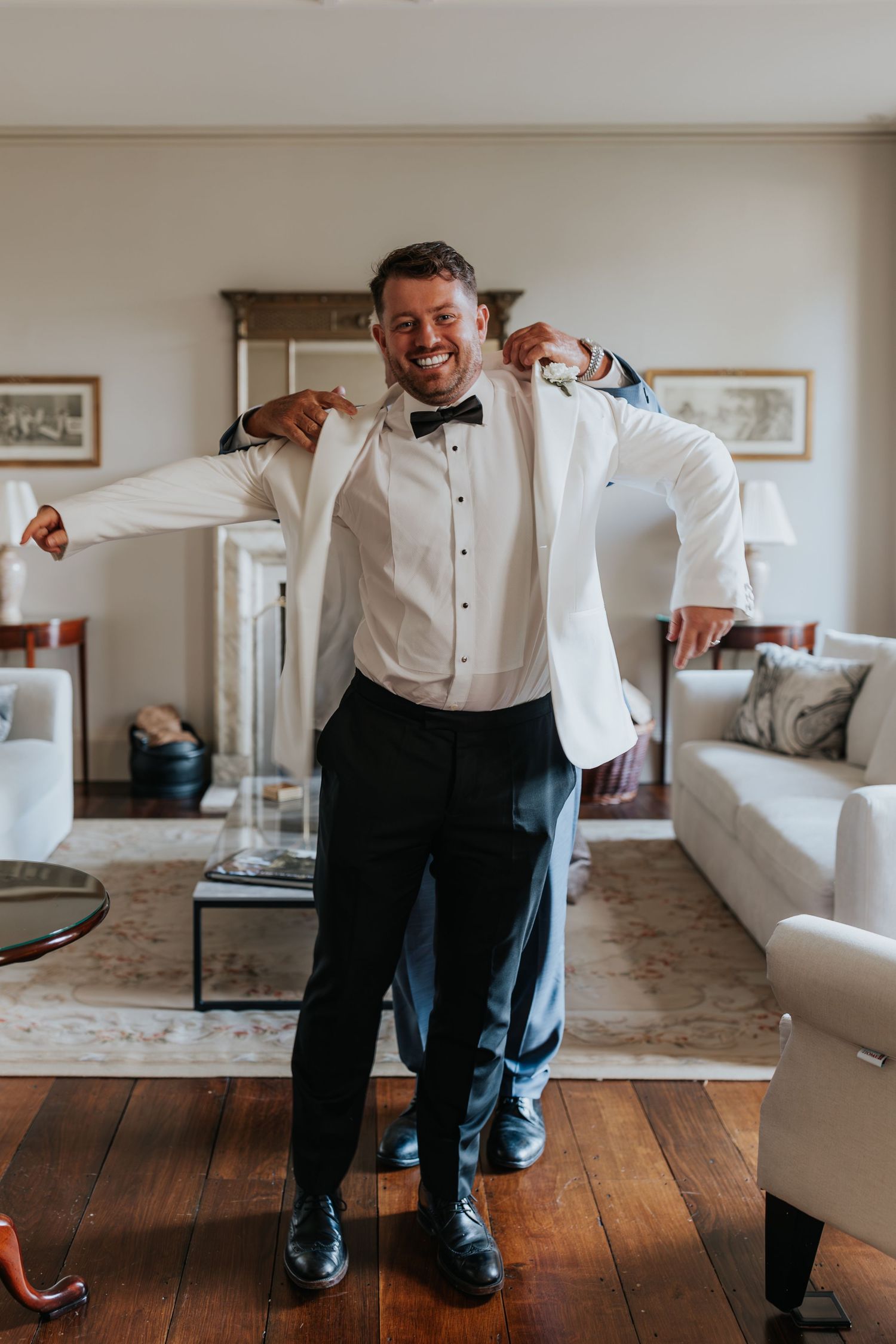 Groom in white tuxedo jacket and black bow tie smiling while getting dressed in elegant hotel room.