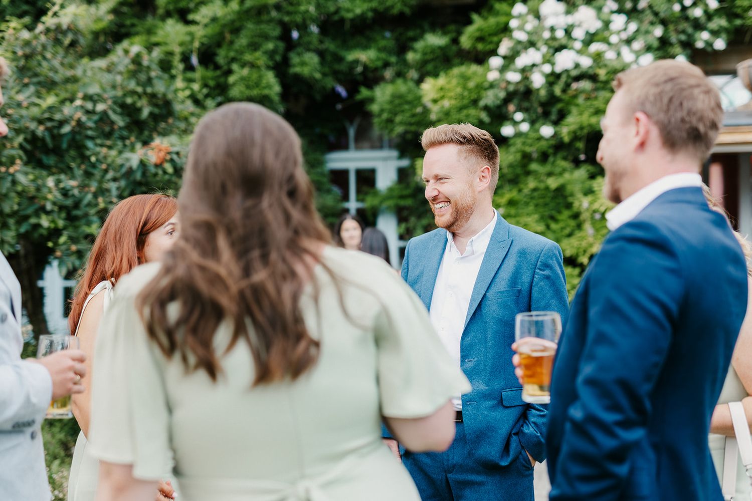 Guests socializing outdoors at a wedding reception, man in blue suit holding a beer glass smiling.