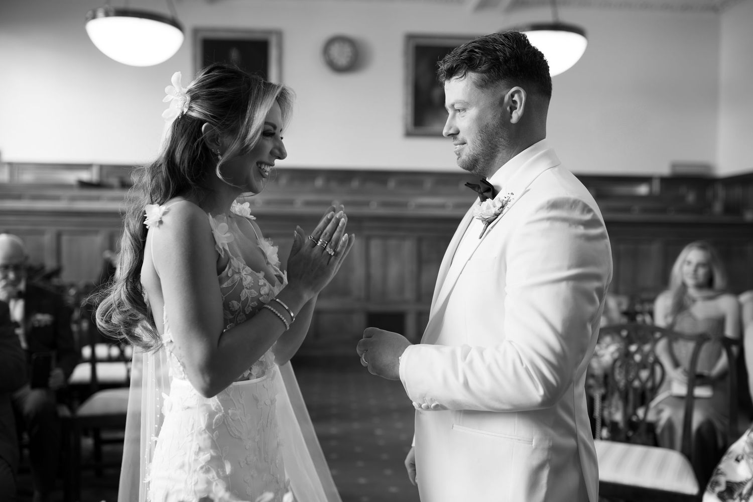 Black and white wedding ceremony photo of bride and groom exchanging vows at elegant indoor venue.