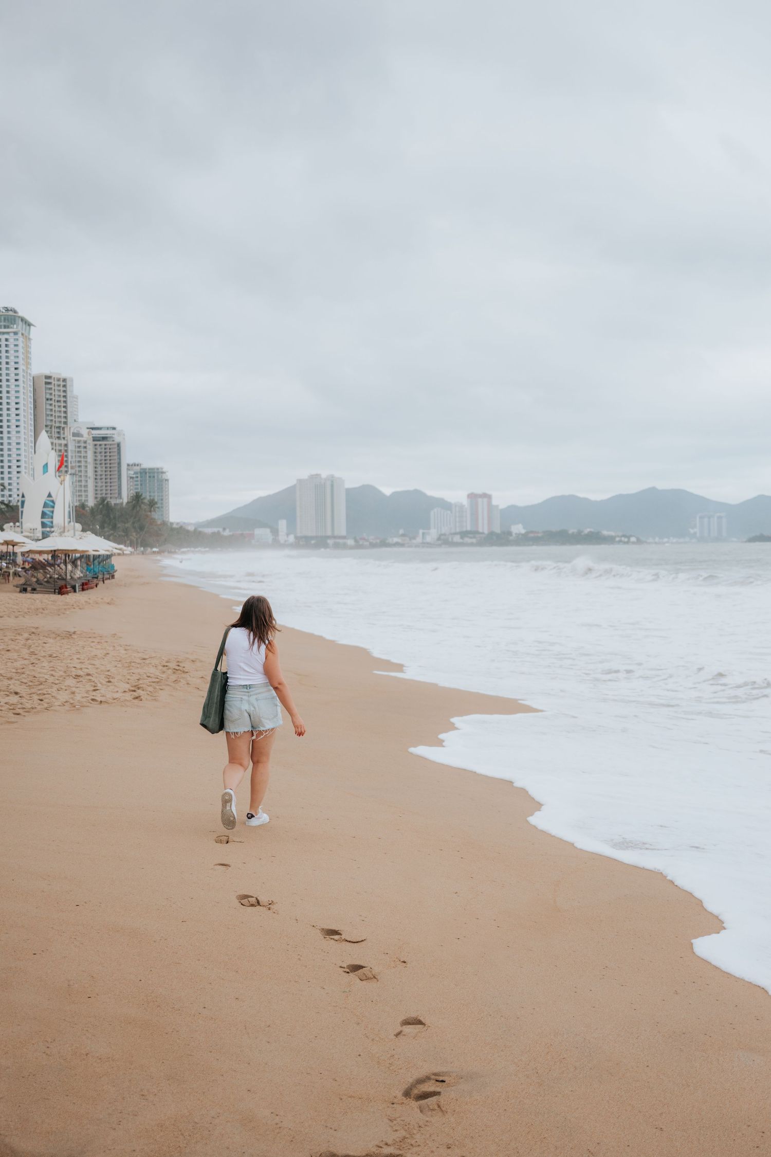 A person walks along a sandy beach with high-rise buildings in the background on a cloudy day.