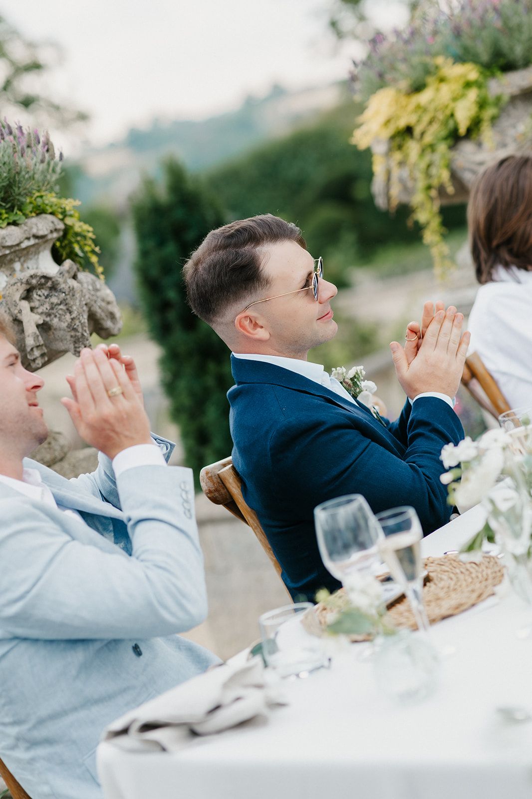 Two grooms in suits clapping at an elegant outdoor wedding reception table adorned with floral arrangements and glassware.