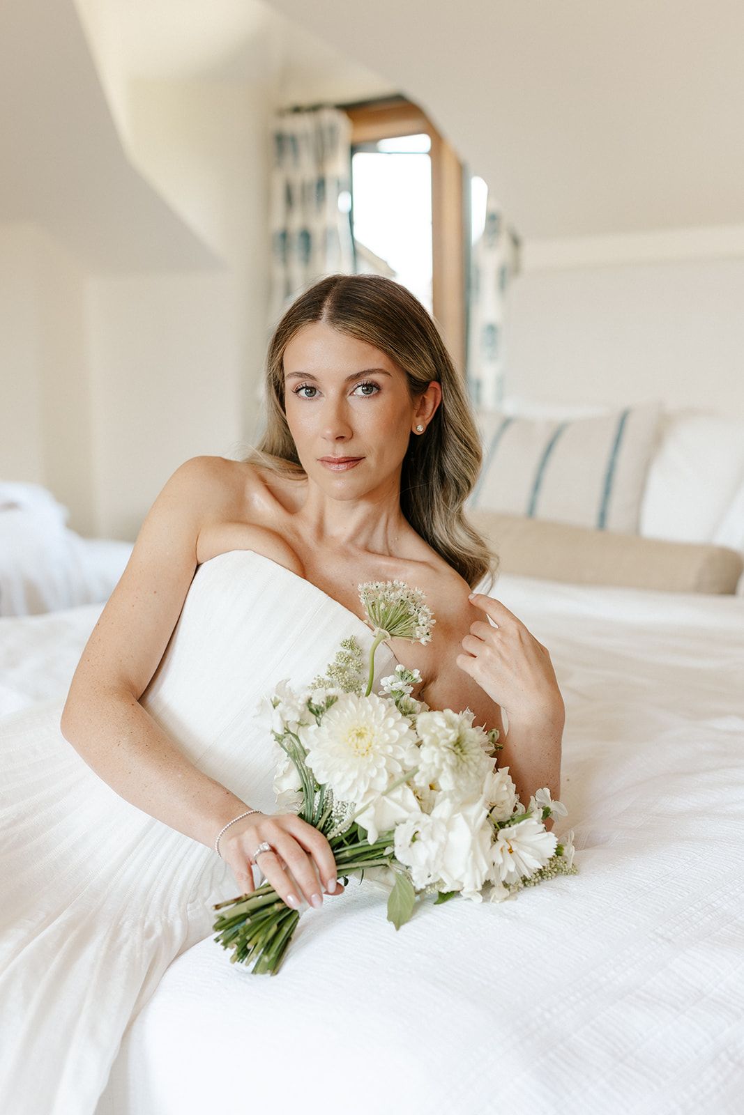 A bride in a strapless white gown sits on a bed holding a white floral bouquet, gazing softly at the camera.