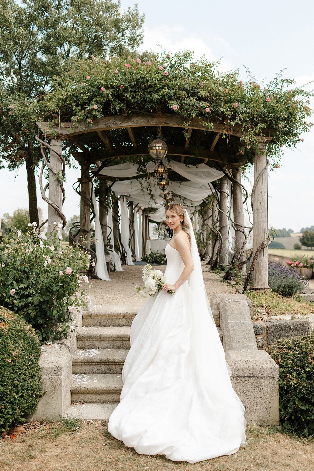 Bride in white ball gown holding bouquet stands before a flower-covered pergola with stone steps in a garden.