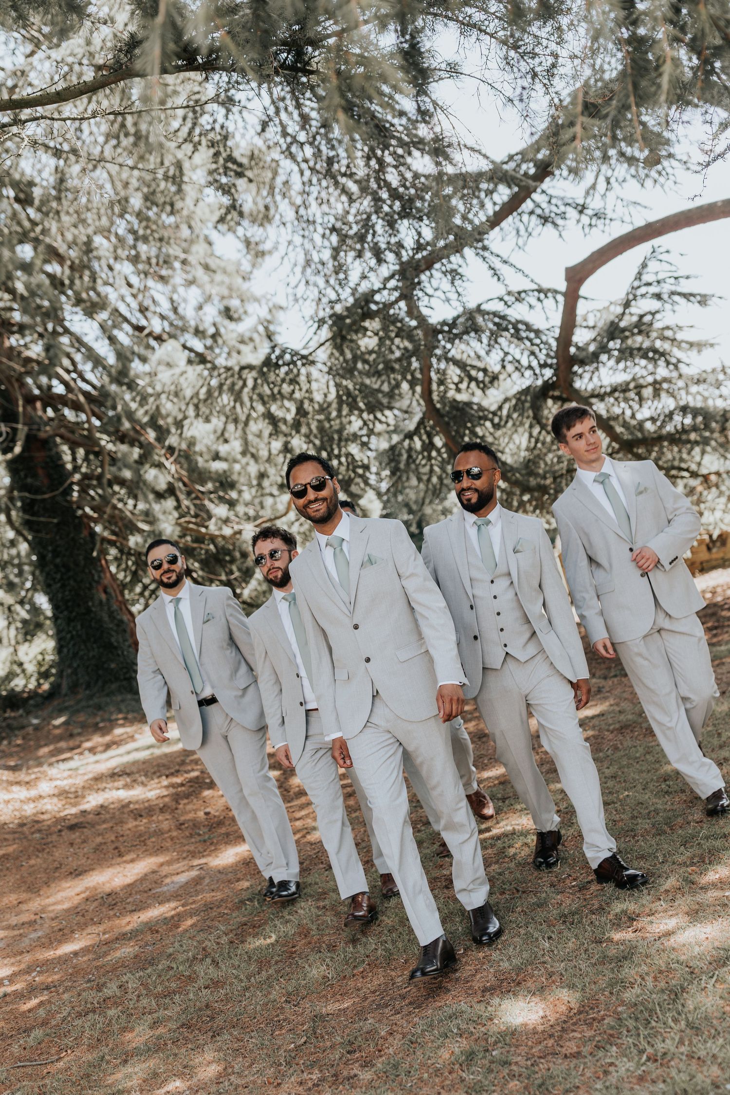 Five groomsmen in light gray suits and sunglasses walk confidently outdoors under a large tree.