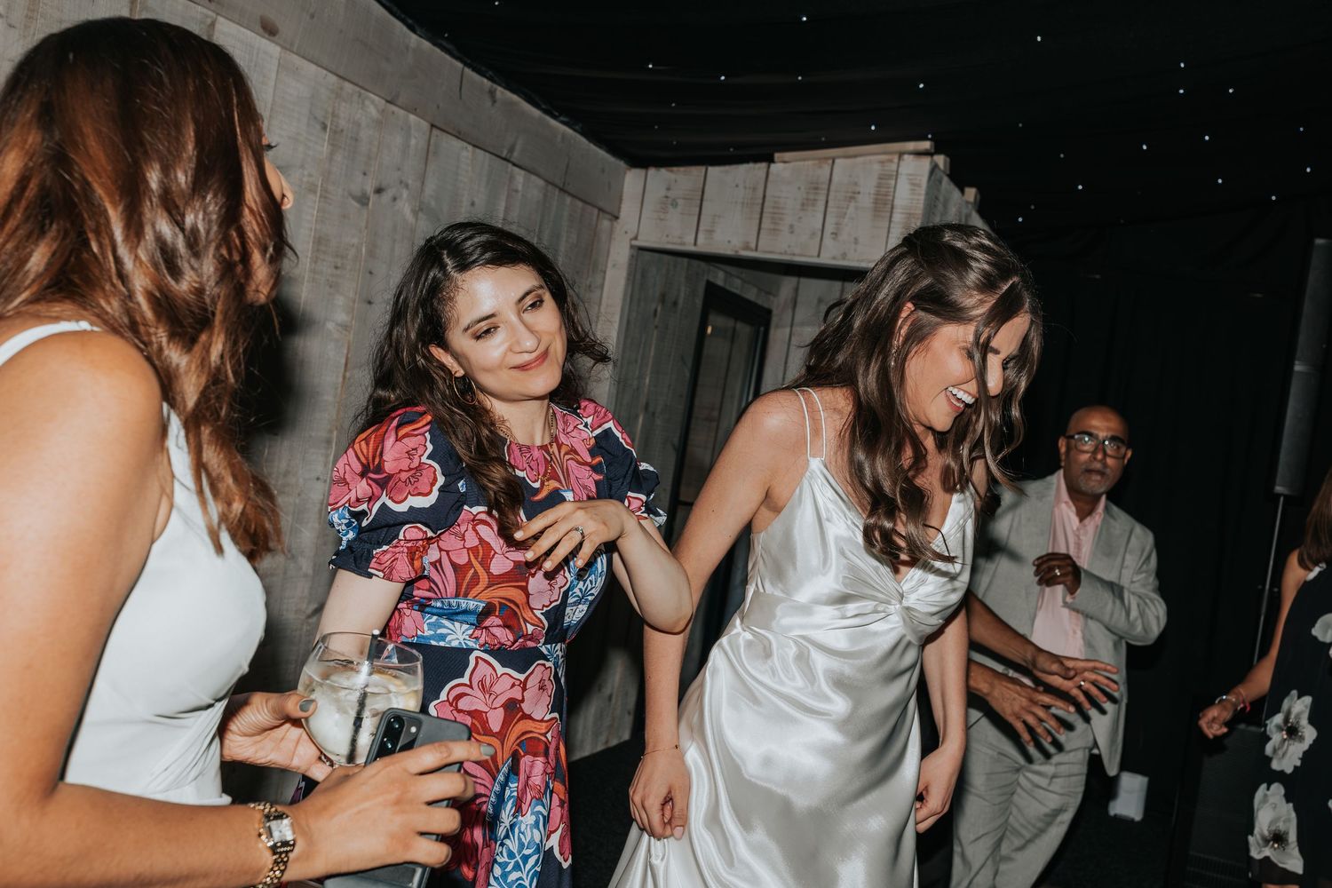 Bride in white satin gown dancing with guests in floral dress at wedding reception celebration.