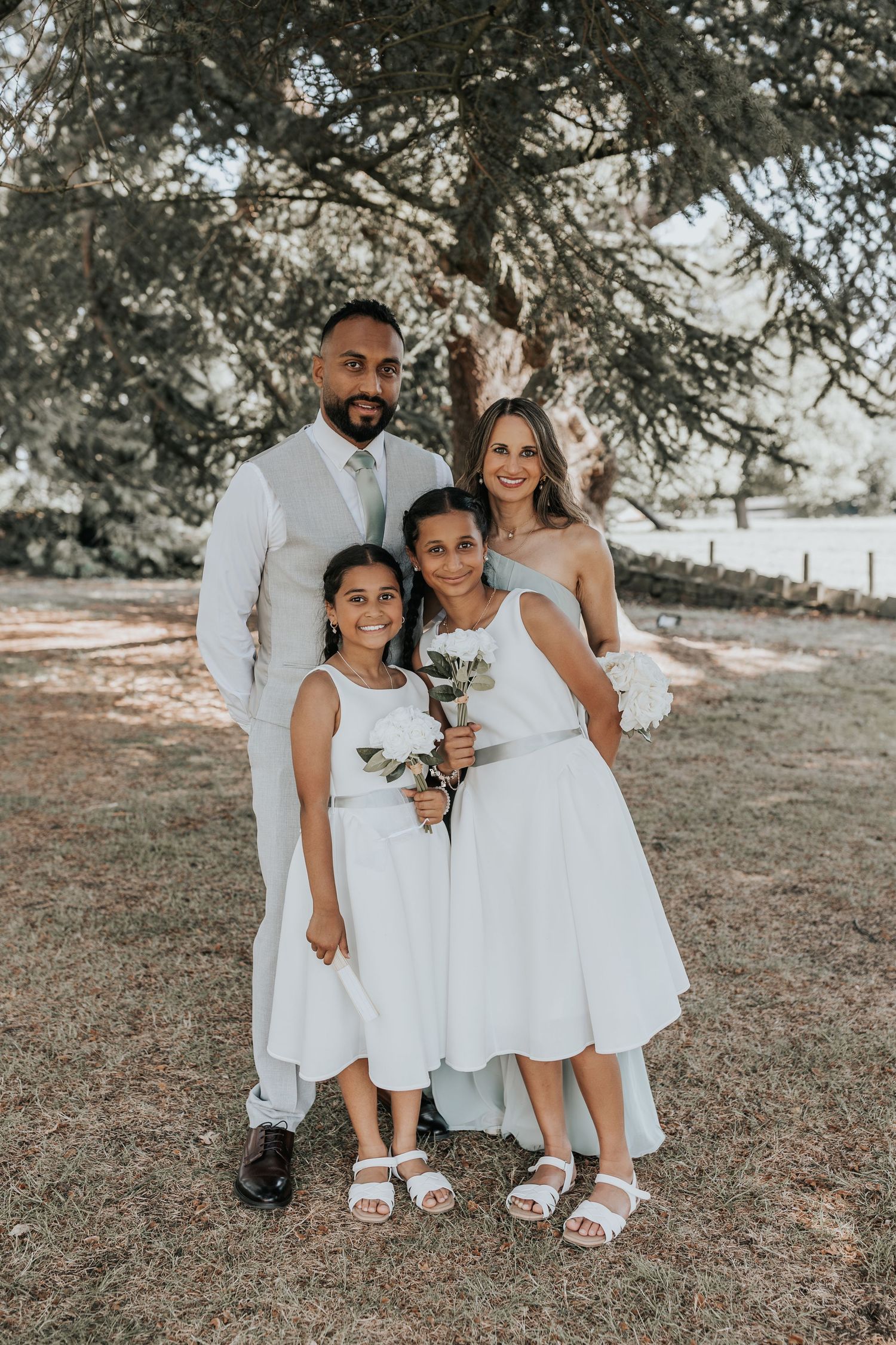 A family of four poses outdoors at a wedding, with the couple in white attire and two girls in white dresses holding flowers.