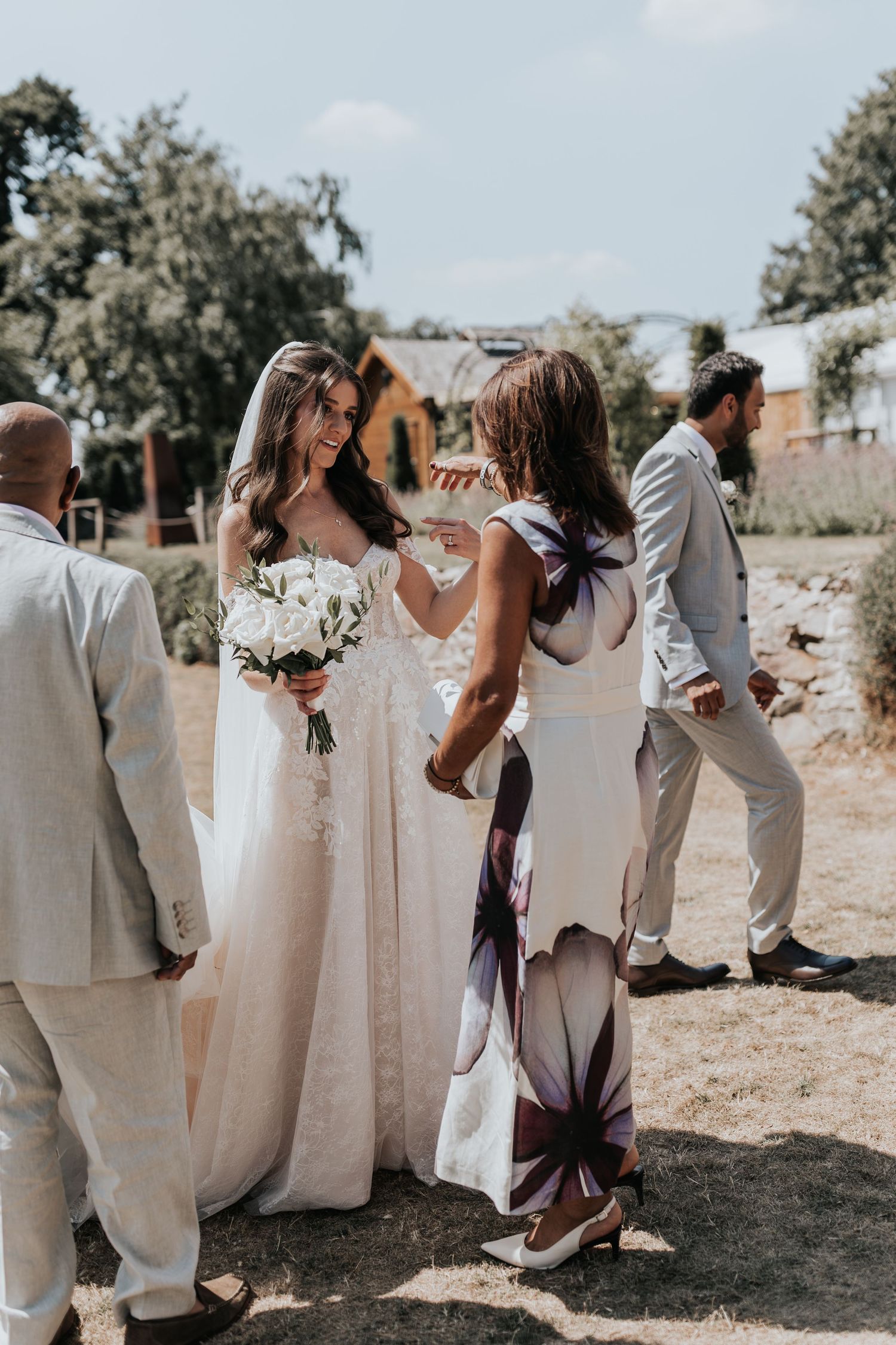 Bride in lace gown holding white bouquet talks with guest in floral dress at outdoor wedding reception.