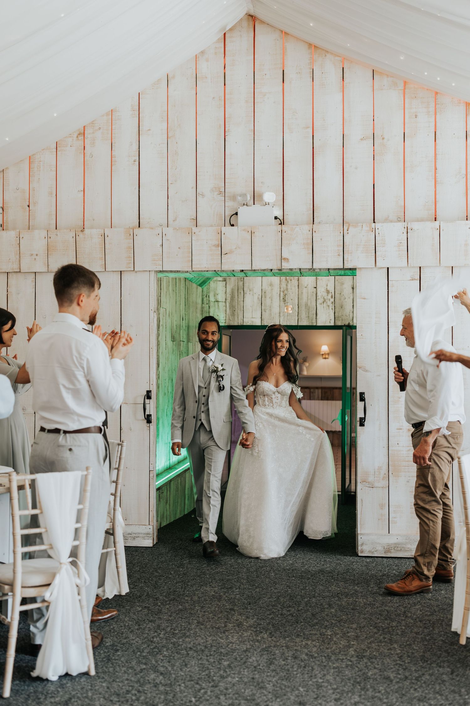 Bride and groom make a grand entrance into a rustic white barn wedding venue as guests applaud.