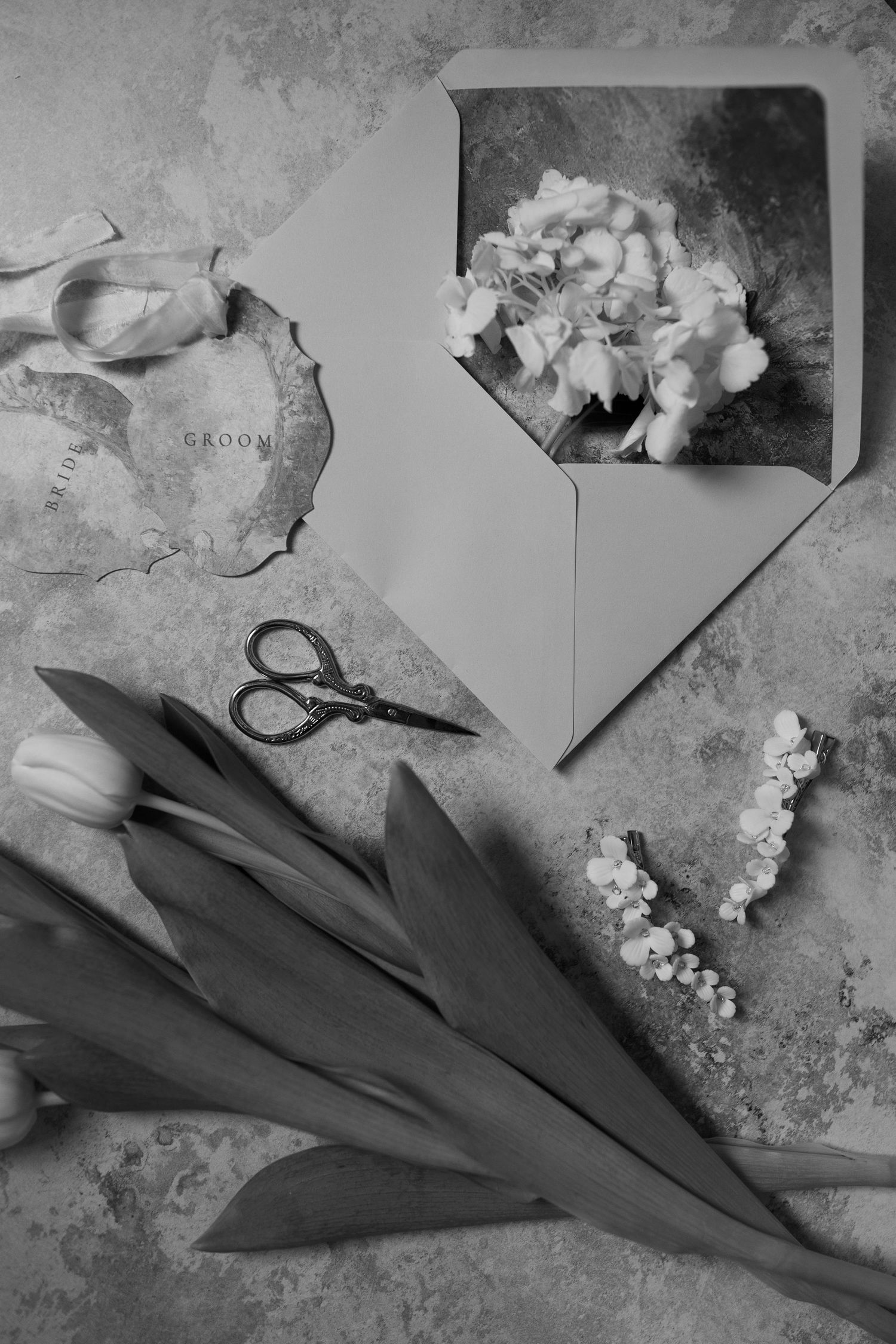 A black and white image showing a floral arrangement, scissors, and an envelope on a textured surface.