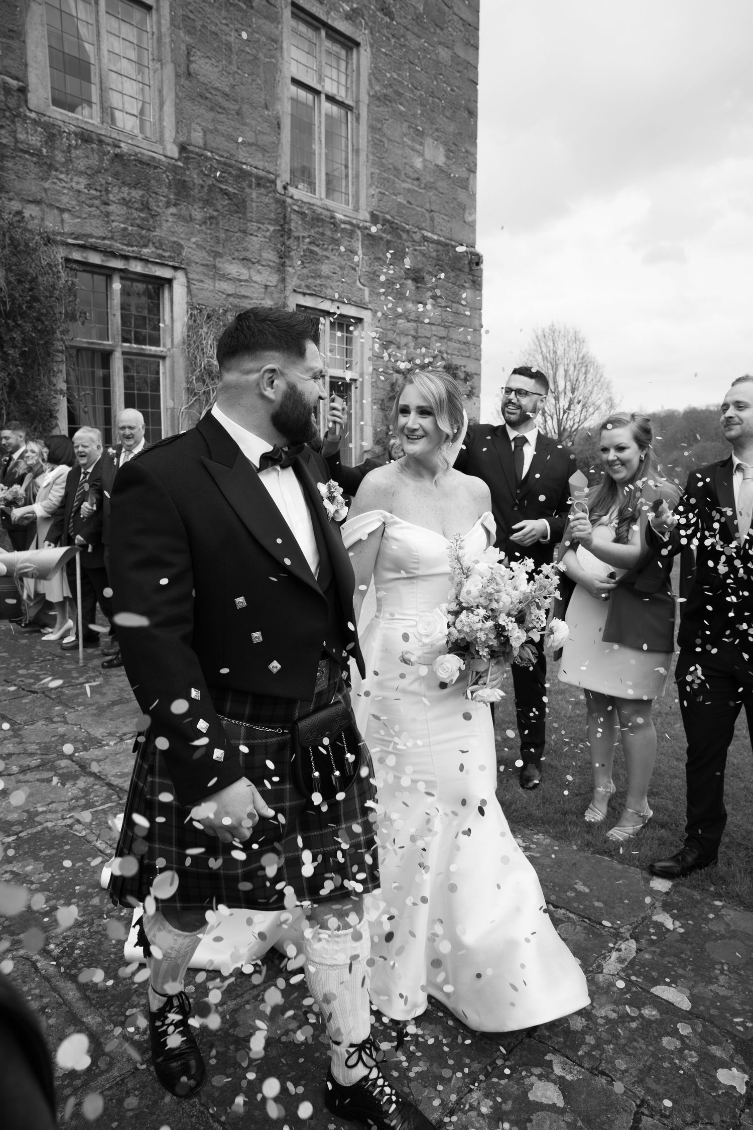 A black and white image of a bride and groom walking together, surrounded by guests throwing confetti at them in Lake District
