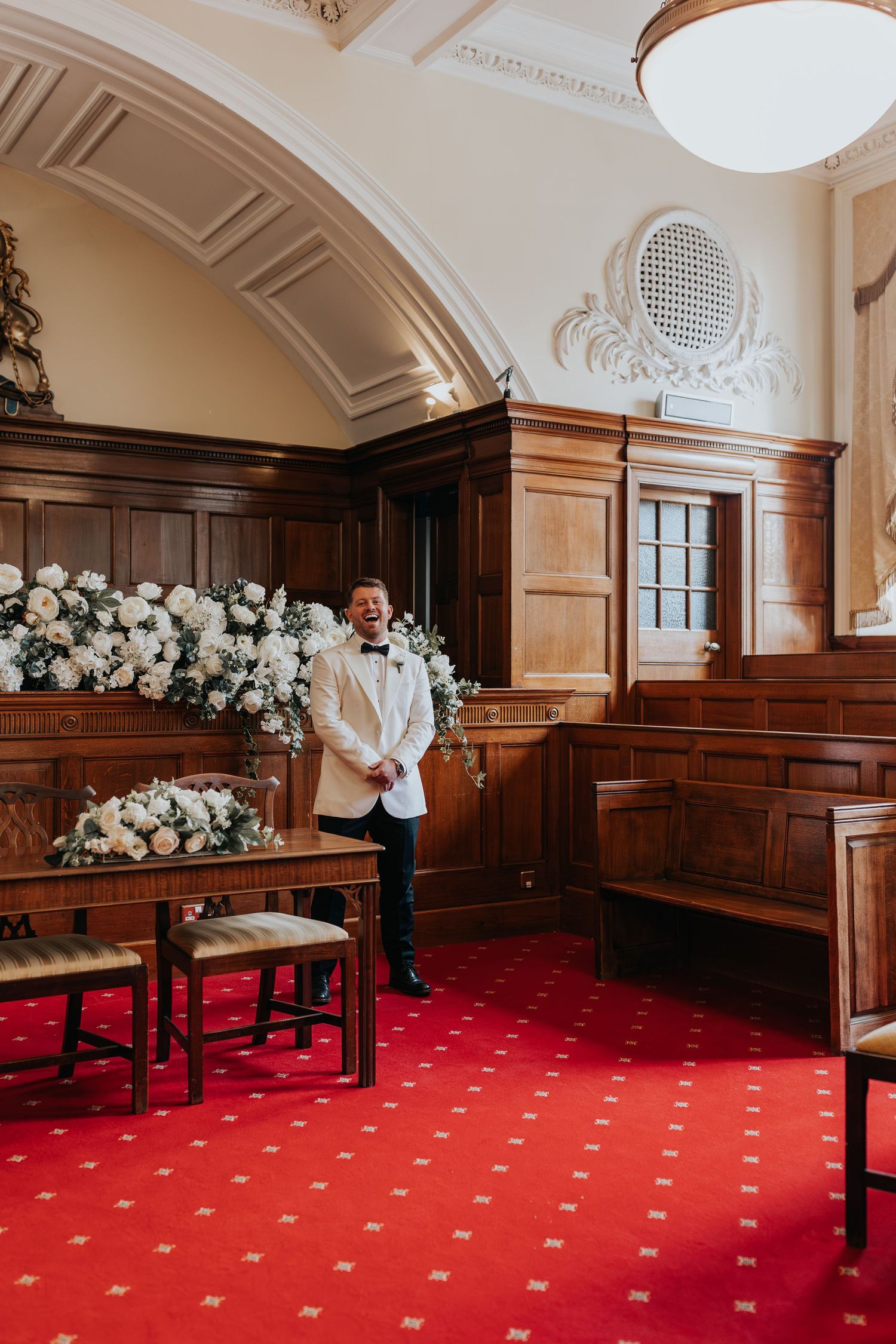 Groom in white tuxedo jacket smiling at wedding ceremony venue with red carpet and white floral arrangements.