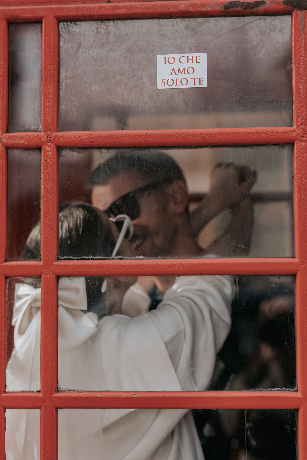 A red telephone booth with a sign that says 'Io che amo solo te', and a couple embracing inside the booth.