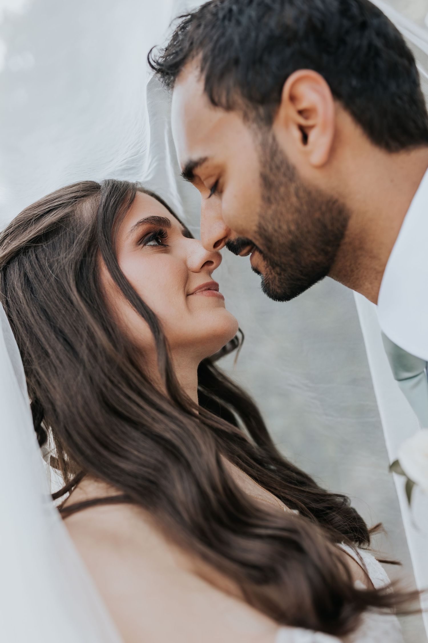 Bride and groom share a romantic close moment under a veil, noses touching and smiling softly.
