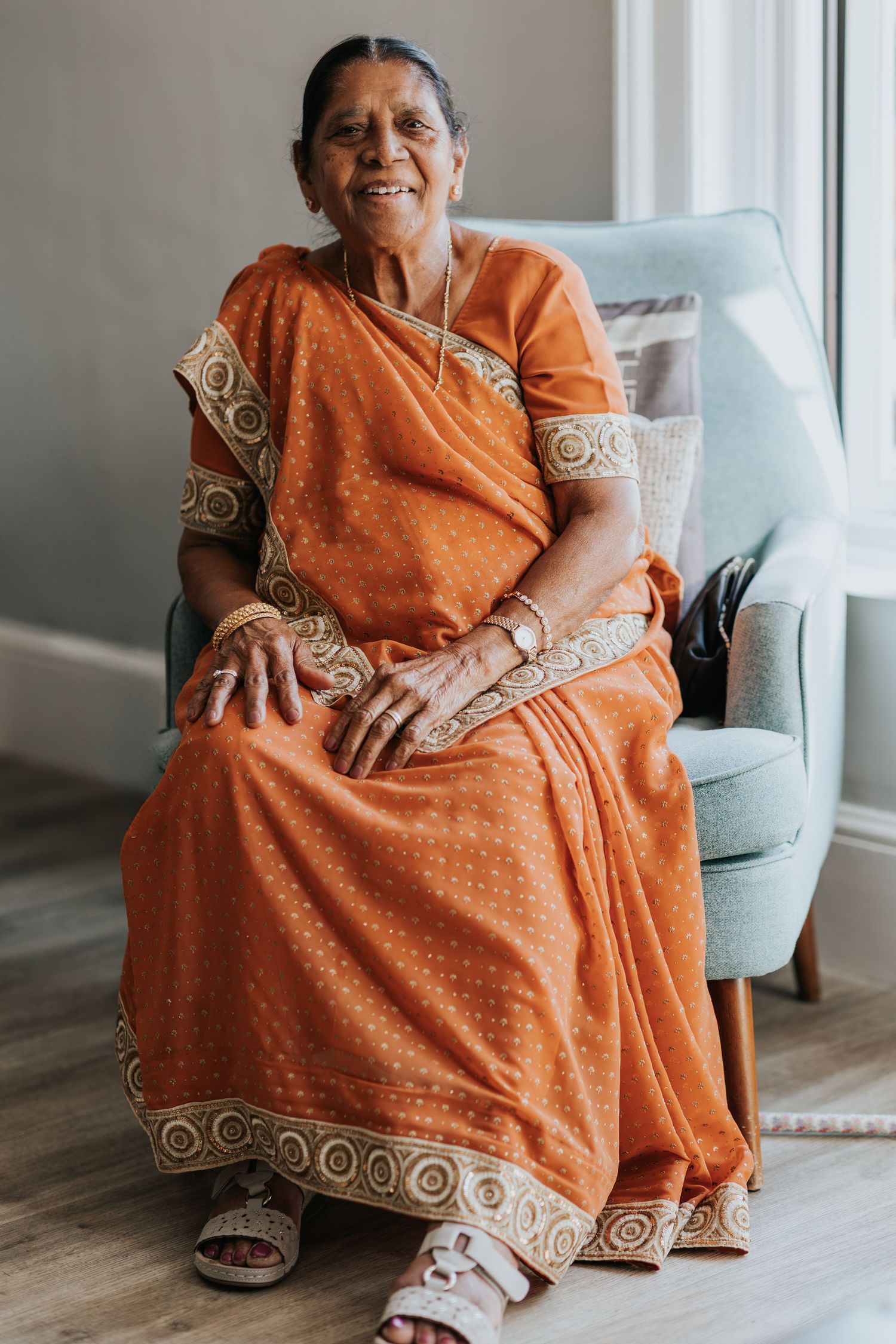 Elderly Indian woman smiling in orange saree with gold trim, seated in light blue chair indoors.