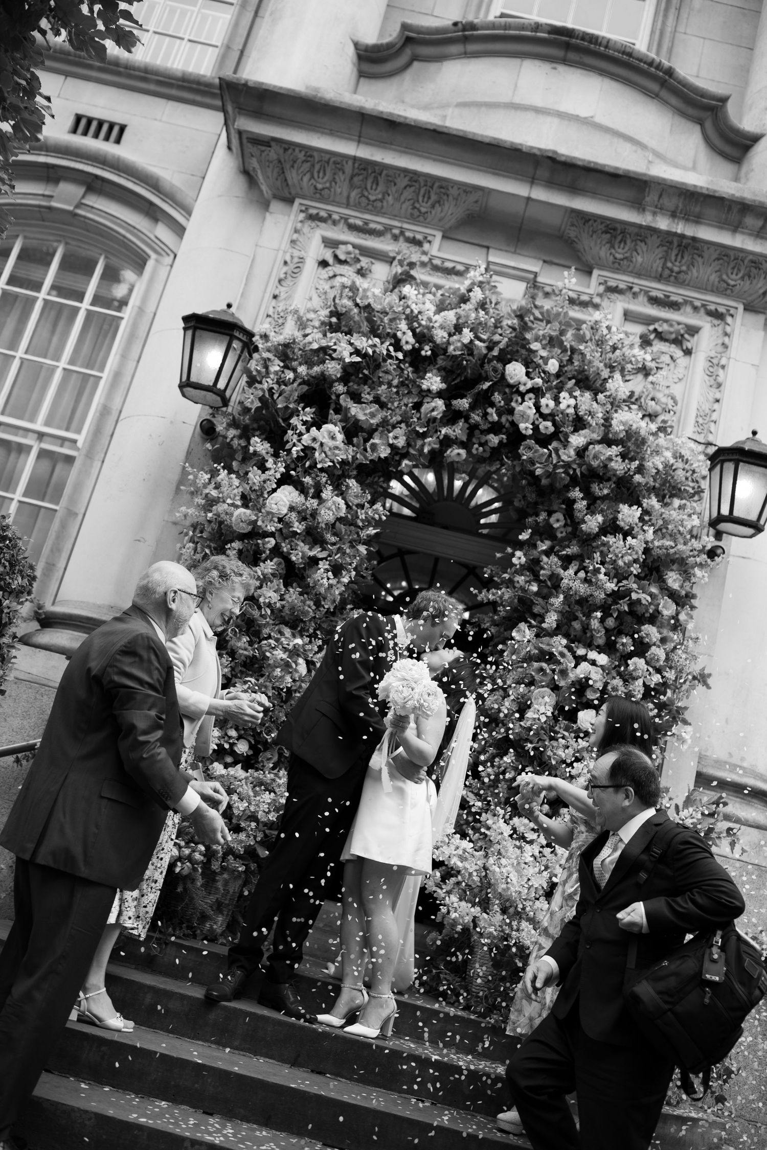 A black and white photograph of a wedding party exiting a building decorated with flowers.