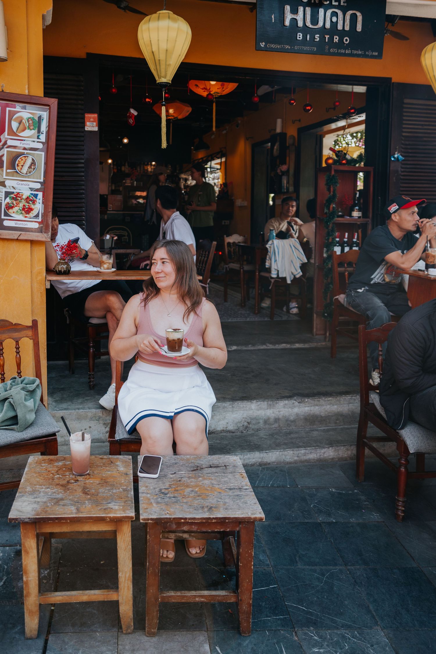 A woman sits at an outdoor cafe, enjoying a drink and taking in the lively atmosphere of the surrounding area.