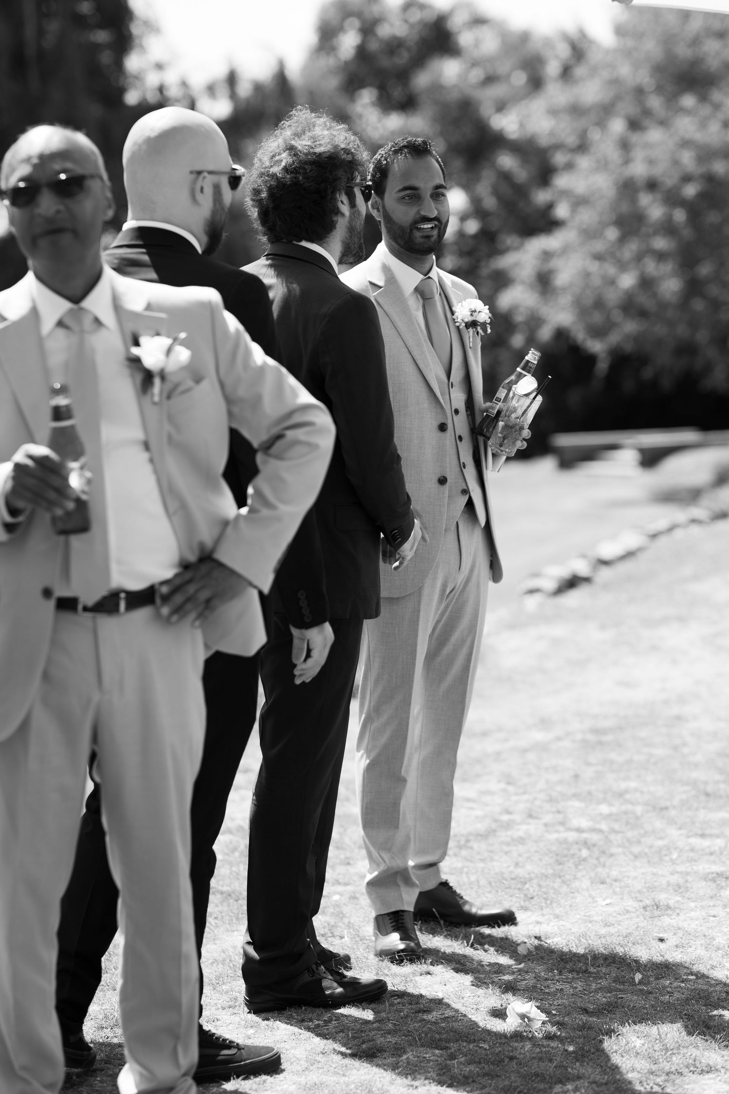 Black and white photo of groomsmen in suits socializing outdoors at a wedding reception on a sunny day.