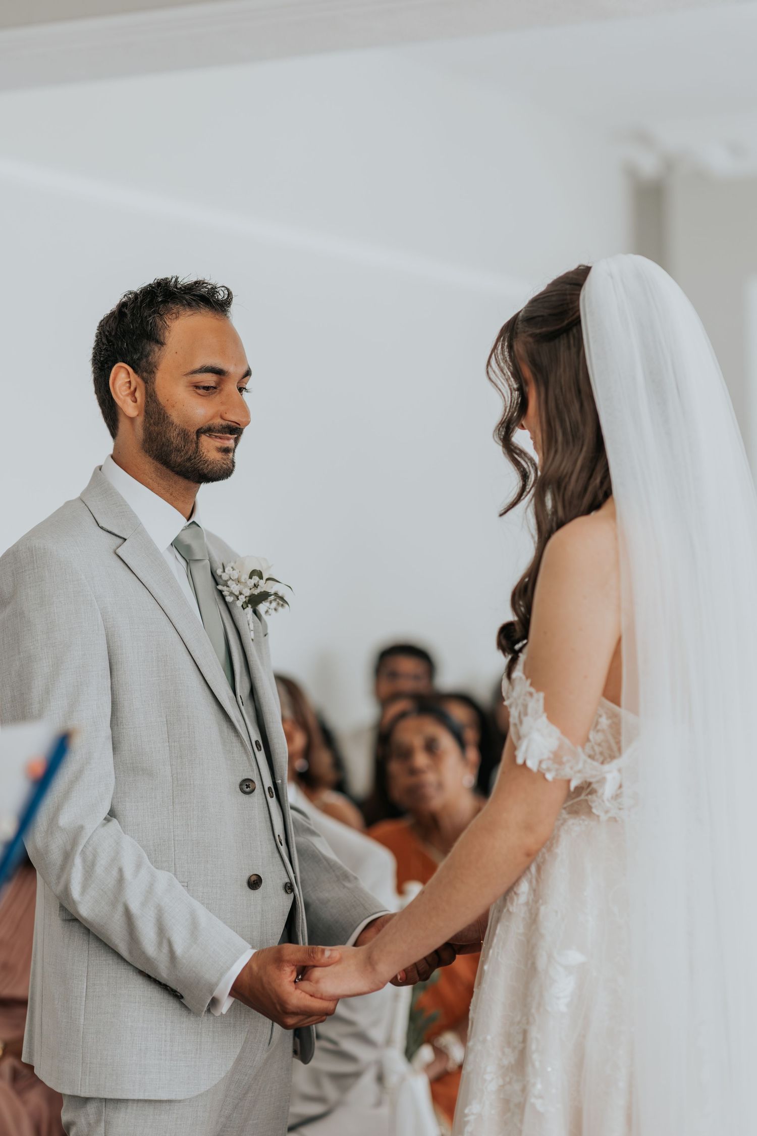 Groom in gray suit smiles at bride in lace wedding dress during ceremony, holding hands at the altar.