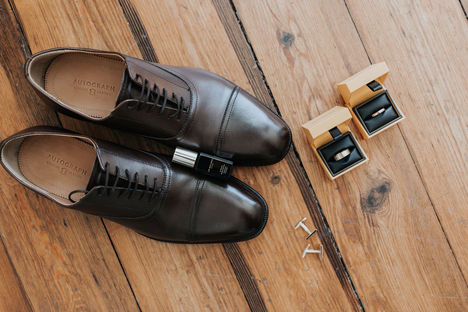 Brown leather oxford dress shoes with black laces beside cufflinks and accessories on rustic wooden floor.