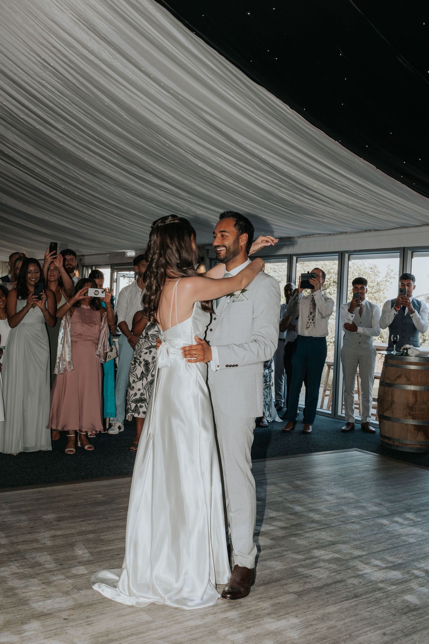 Bride and groom share their first dance surrounded by cheering guests in elegant reception venue with draped ceiling.