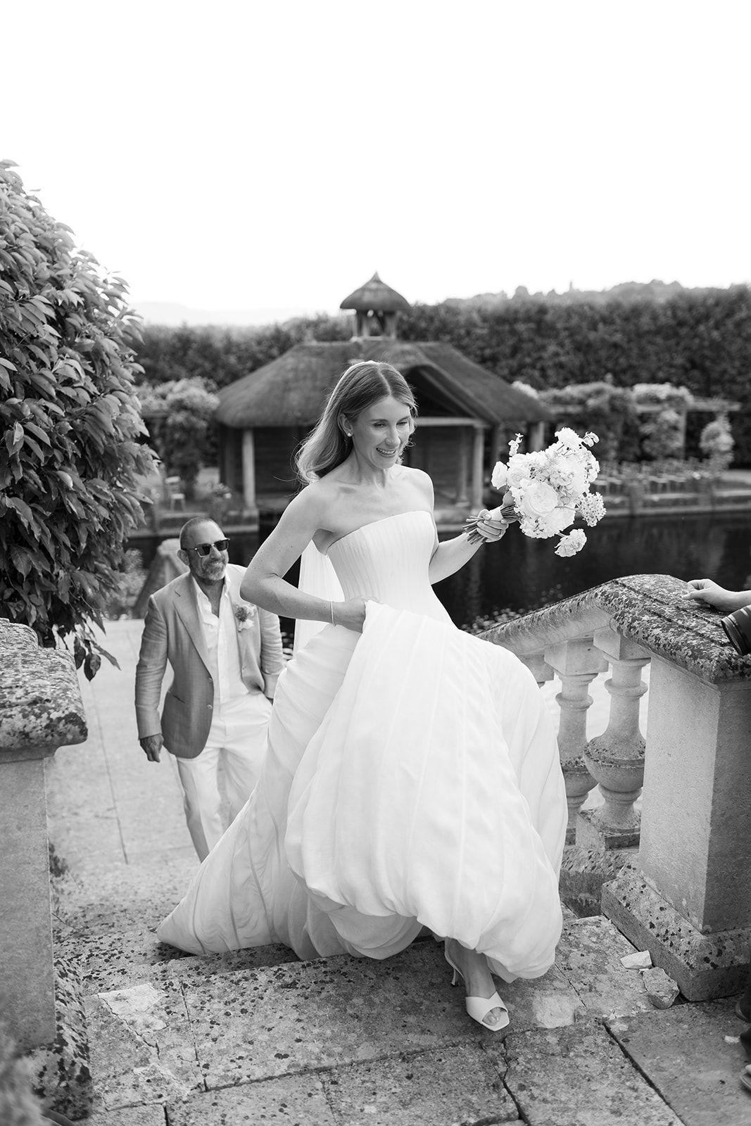 Black and white photo of a smiling bride in a strapless gown holding a bouquet, with groom following on stone steps.