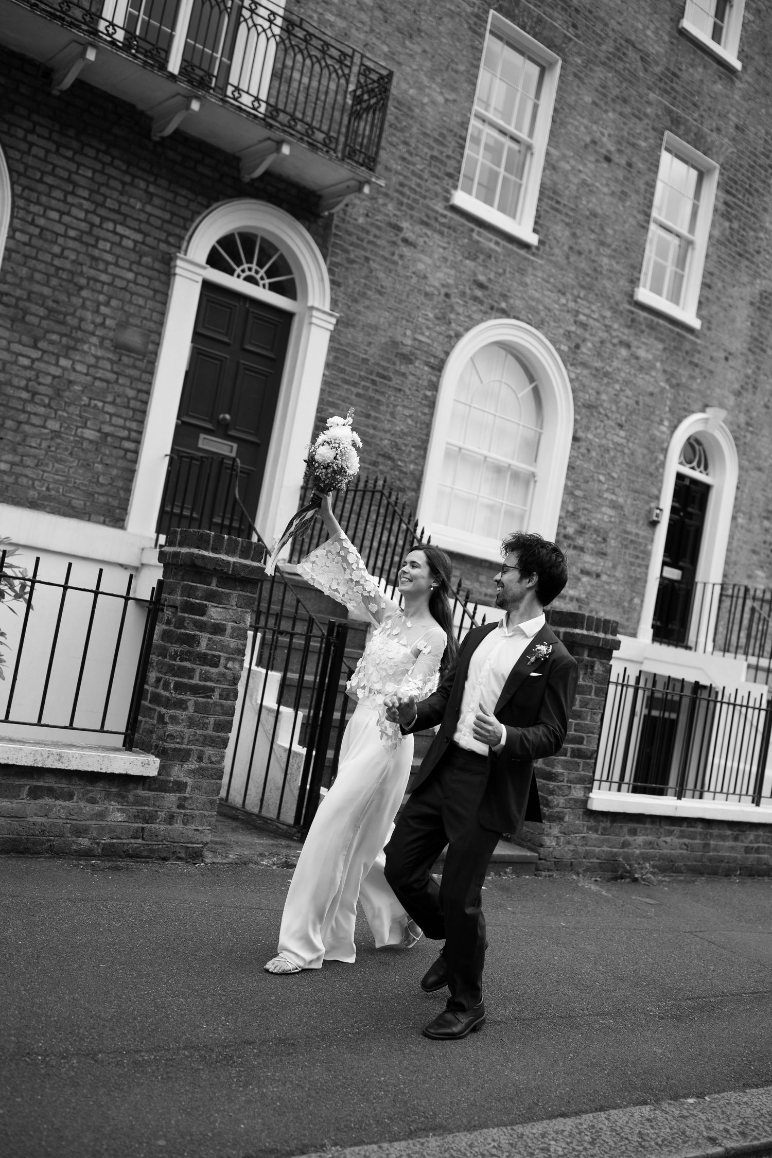 A black and white image of a bride and groom walking together in front of a brick building.