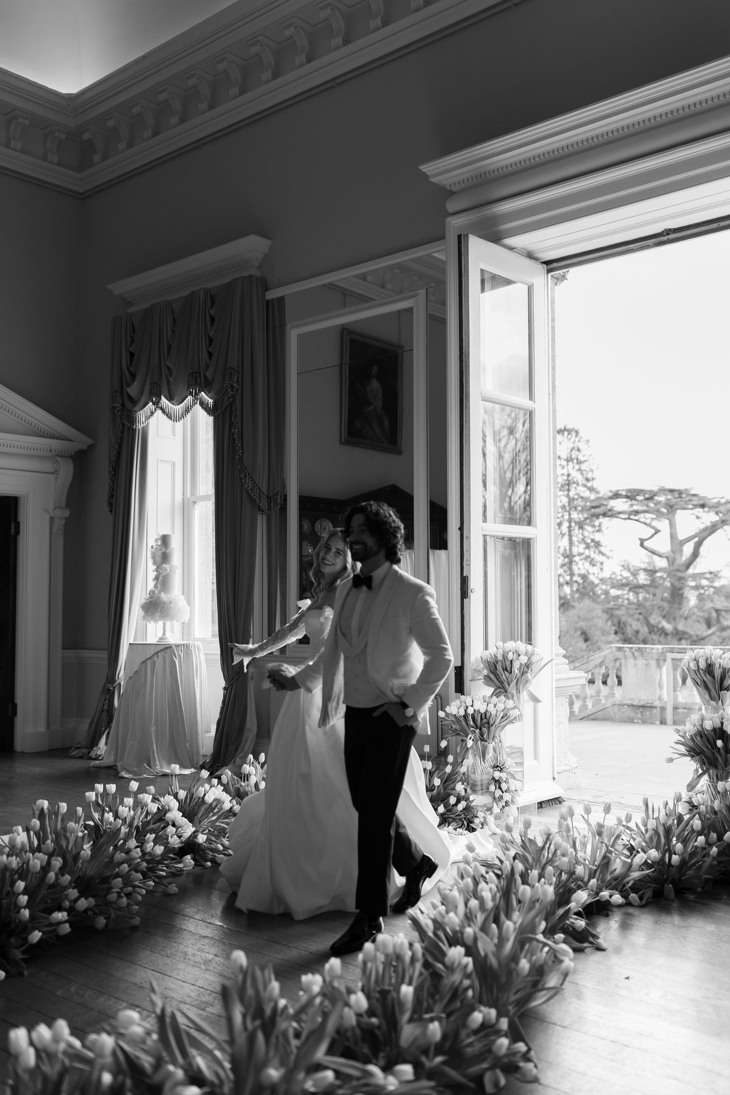 A black and white photograph shows a bride and groom dancing in a room filled with flowers and curtains.
