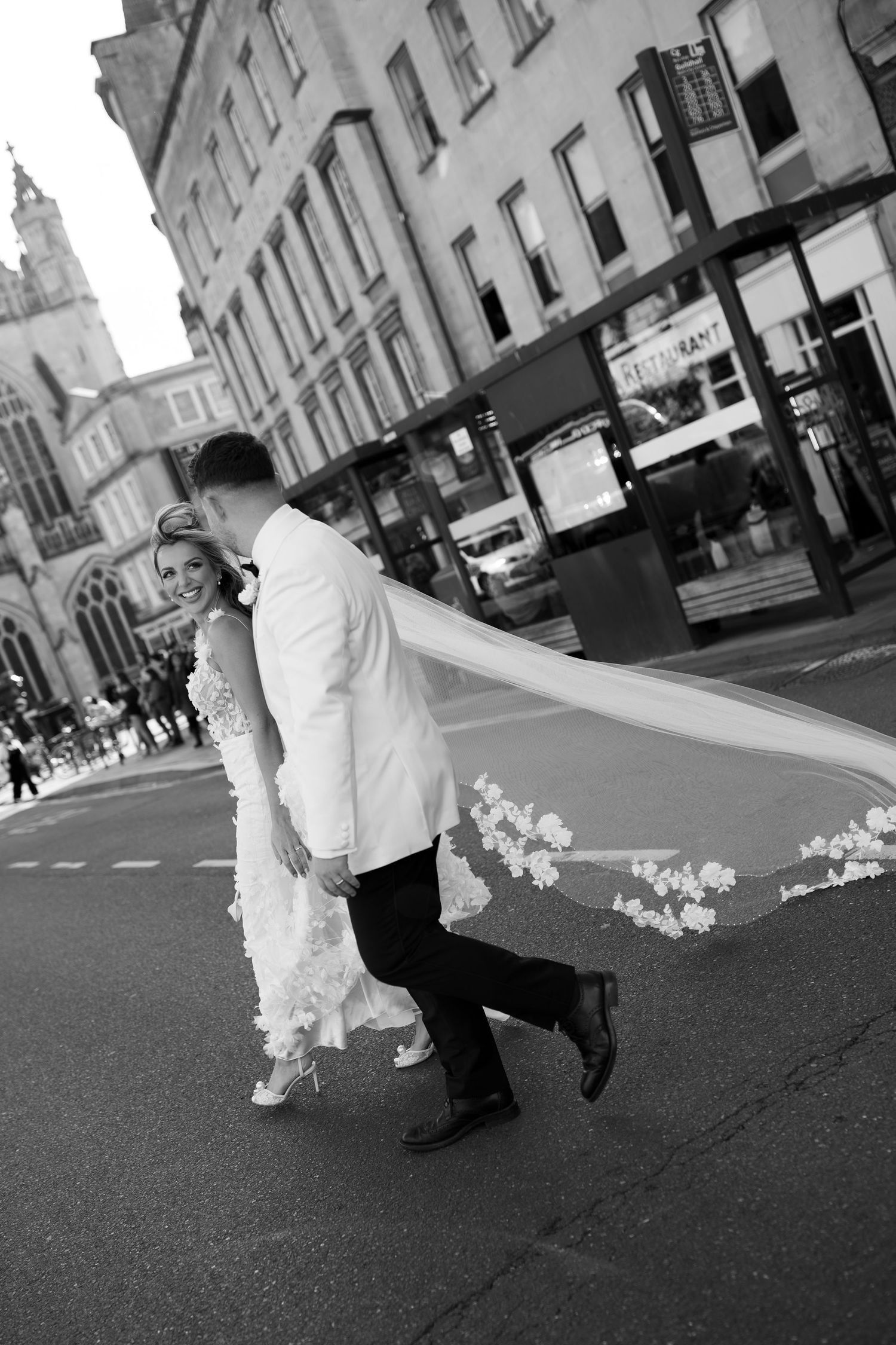 Black and white photo of a bride and groom walking across a city street, her floral veil flowing dramatically in the wind.