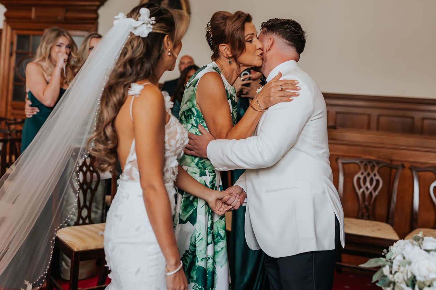 Groom in white suit kissing mother at wedding ceremony while bride in lace gown stands nearby.