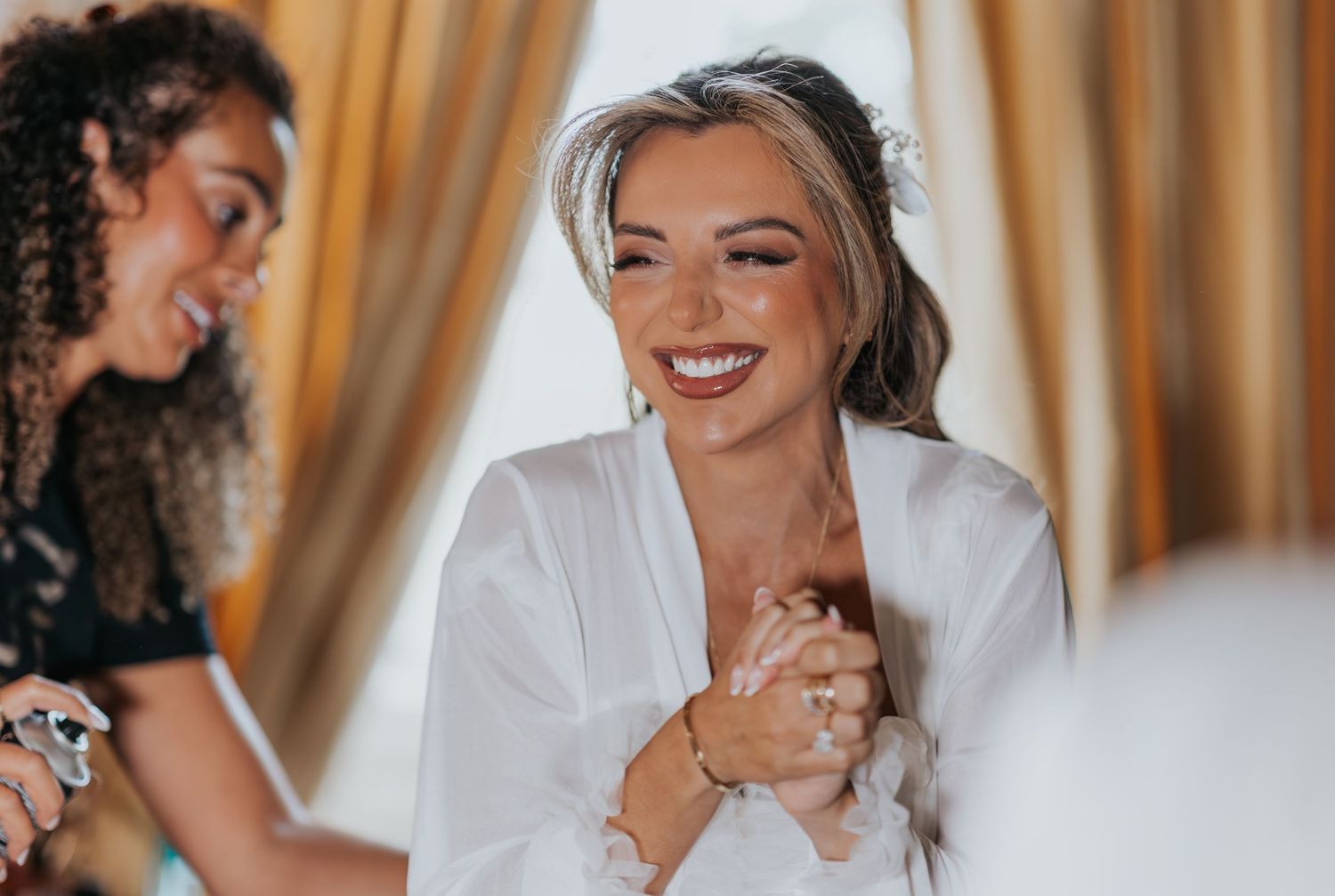 Smiling bride in white robe clasping hands joyfully during wedding preparations with stylist nearby.