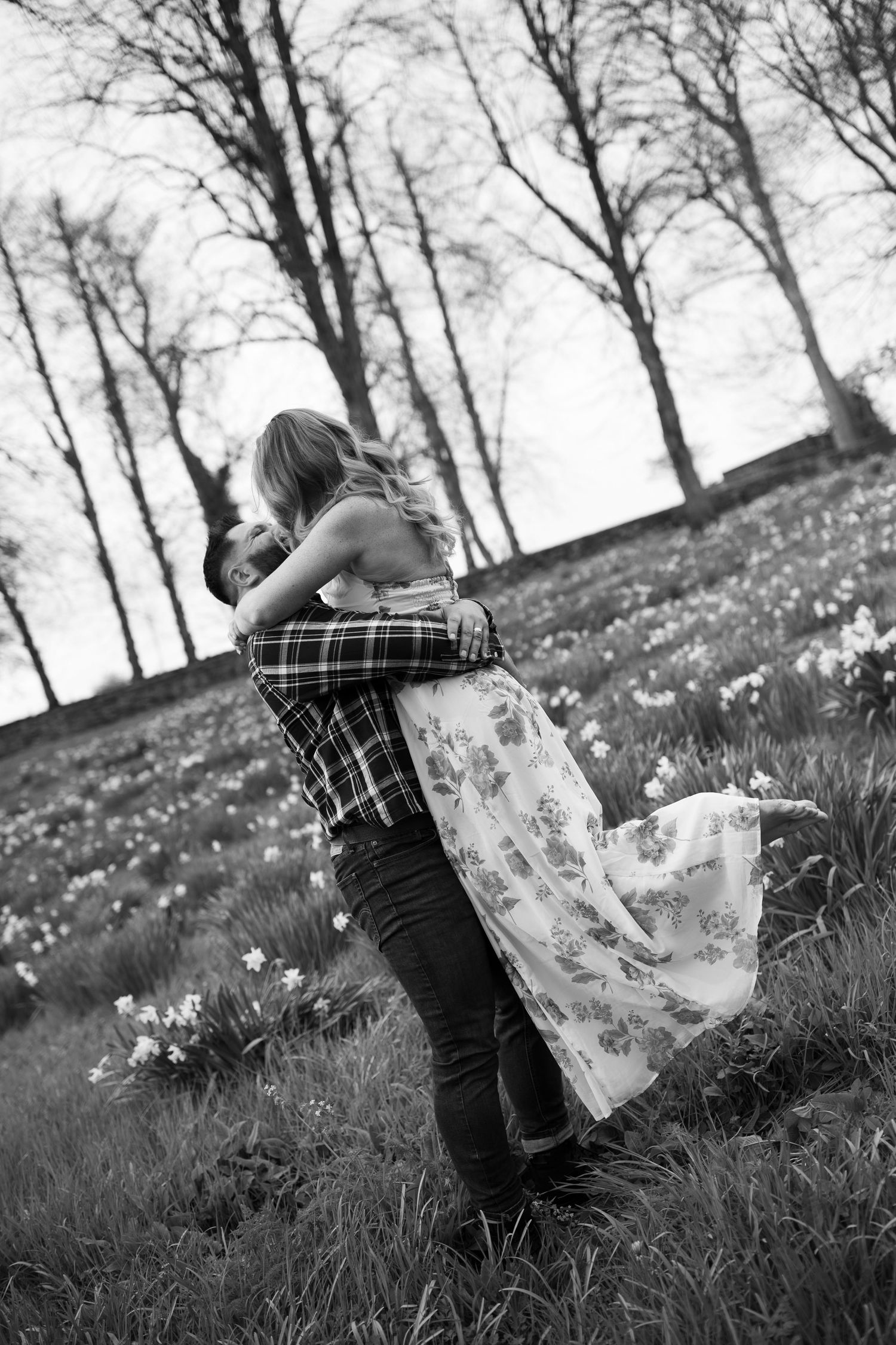 A black and white image shows a couple embracing in a grassy field surrounded by trees.