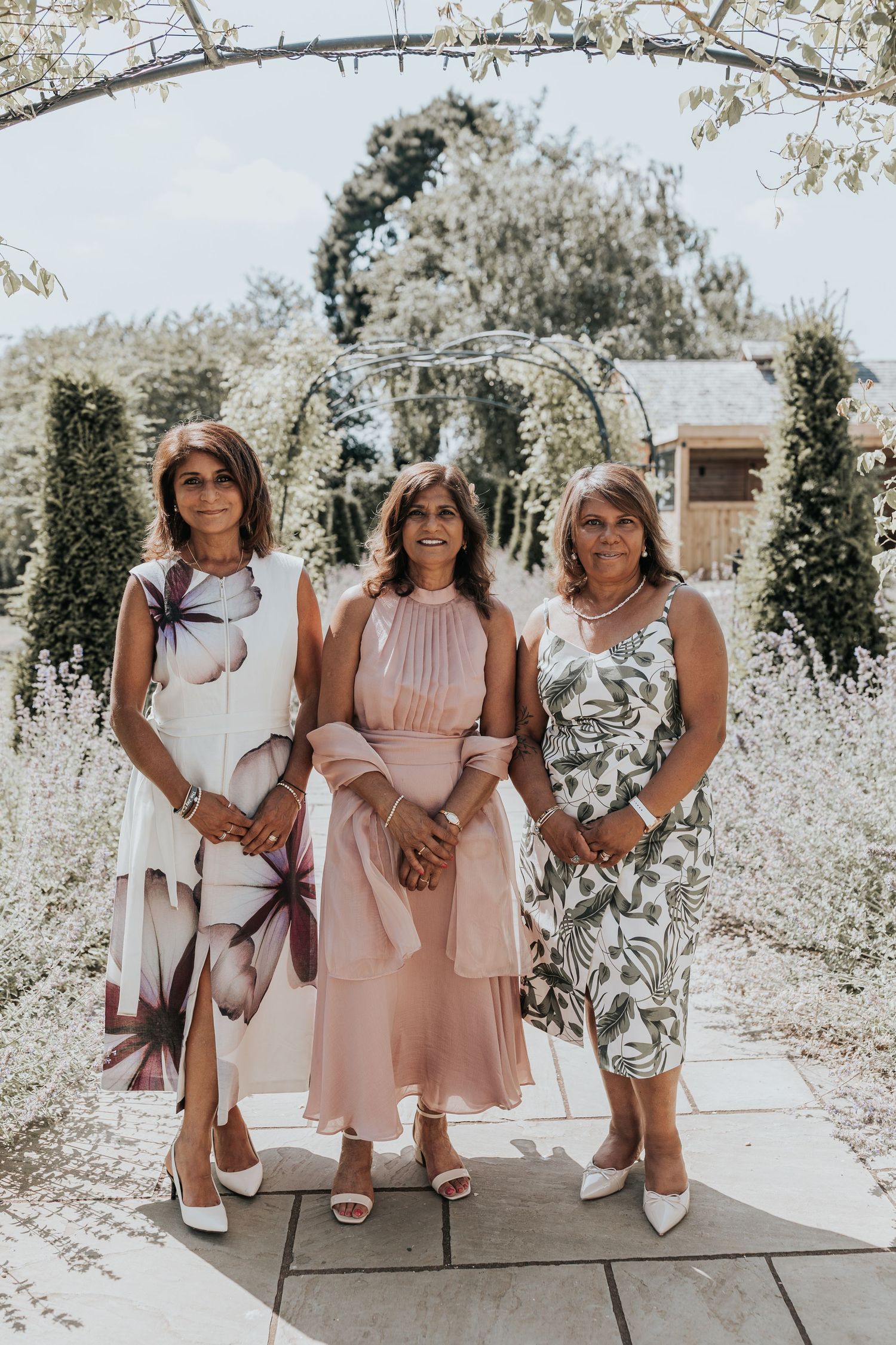 Three women in elegant dresses pose together in a garden with an arched trellis backdrop on a sunny day.