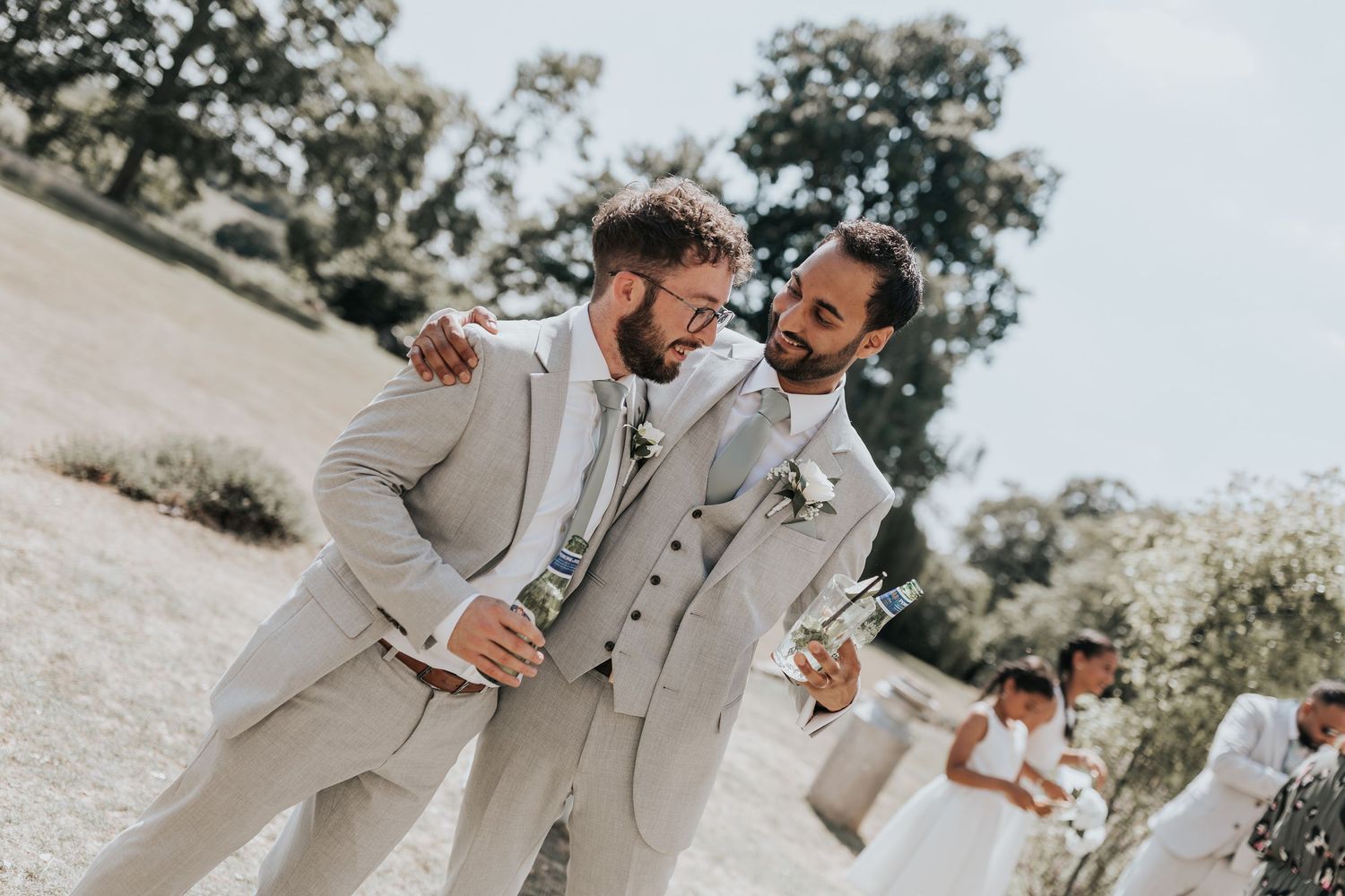 Two grooms in matching beige suits laugh together outdoors at their wedding ceremony on a sunny day.