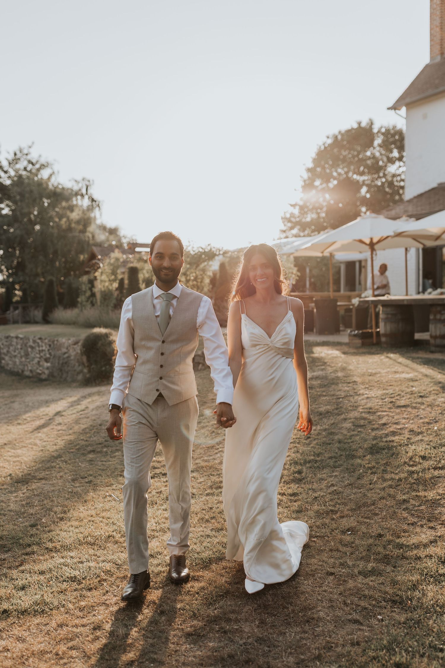 Bride and groom walk hand in hand at golden hour, she in a satin gown, he in a light grey suit.